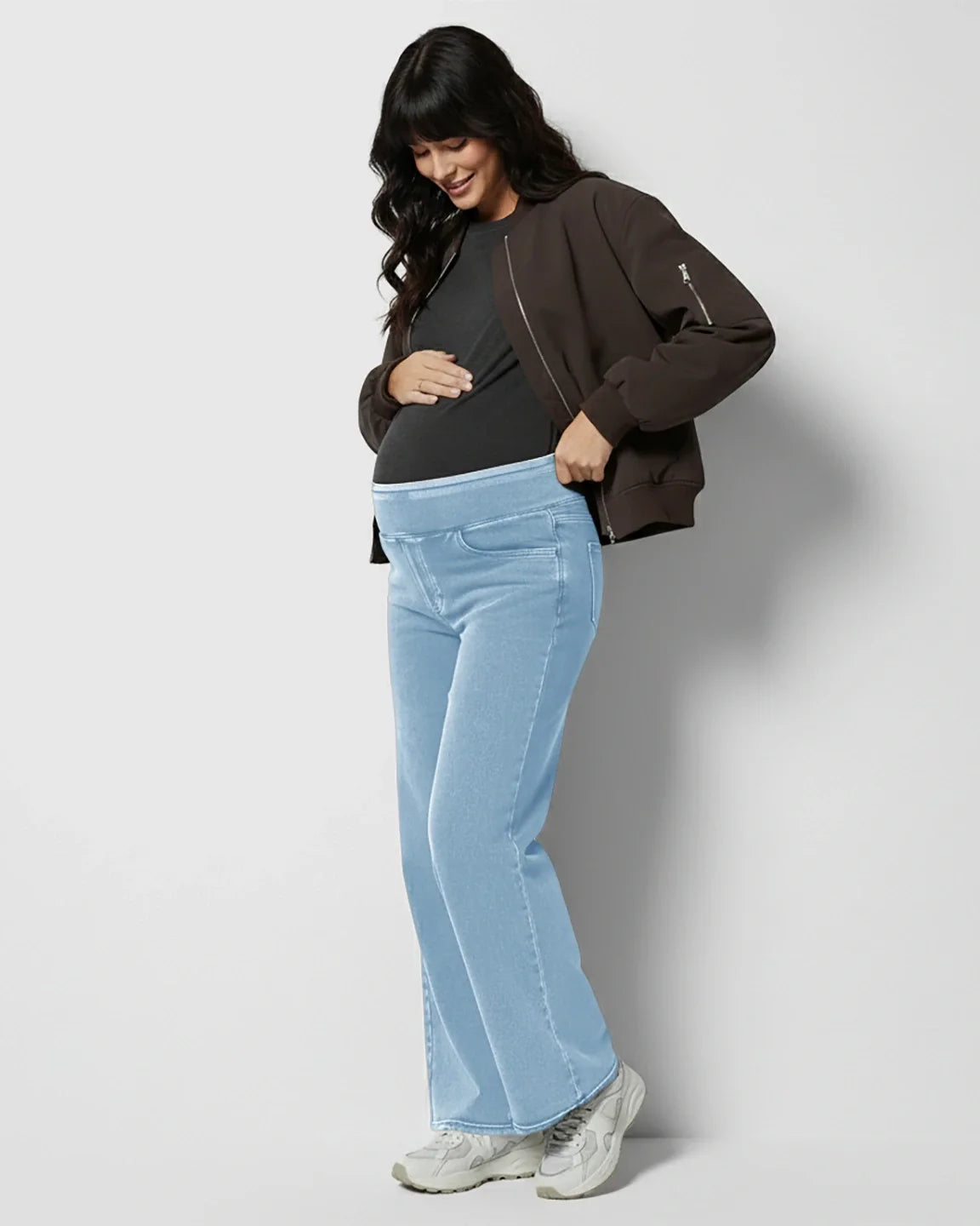 Smiling pregnant woman in light blue wide-leg maternity denim jeans with a soft waistband and a dark bomber jacket.