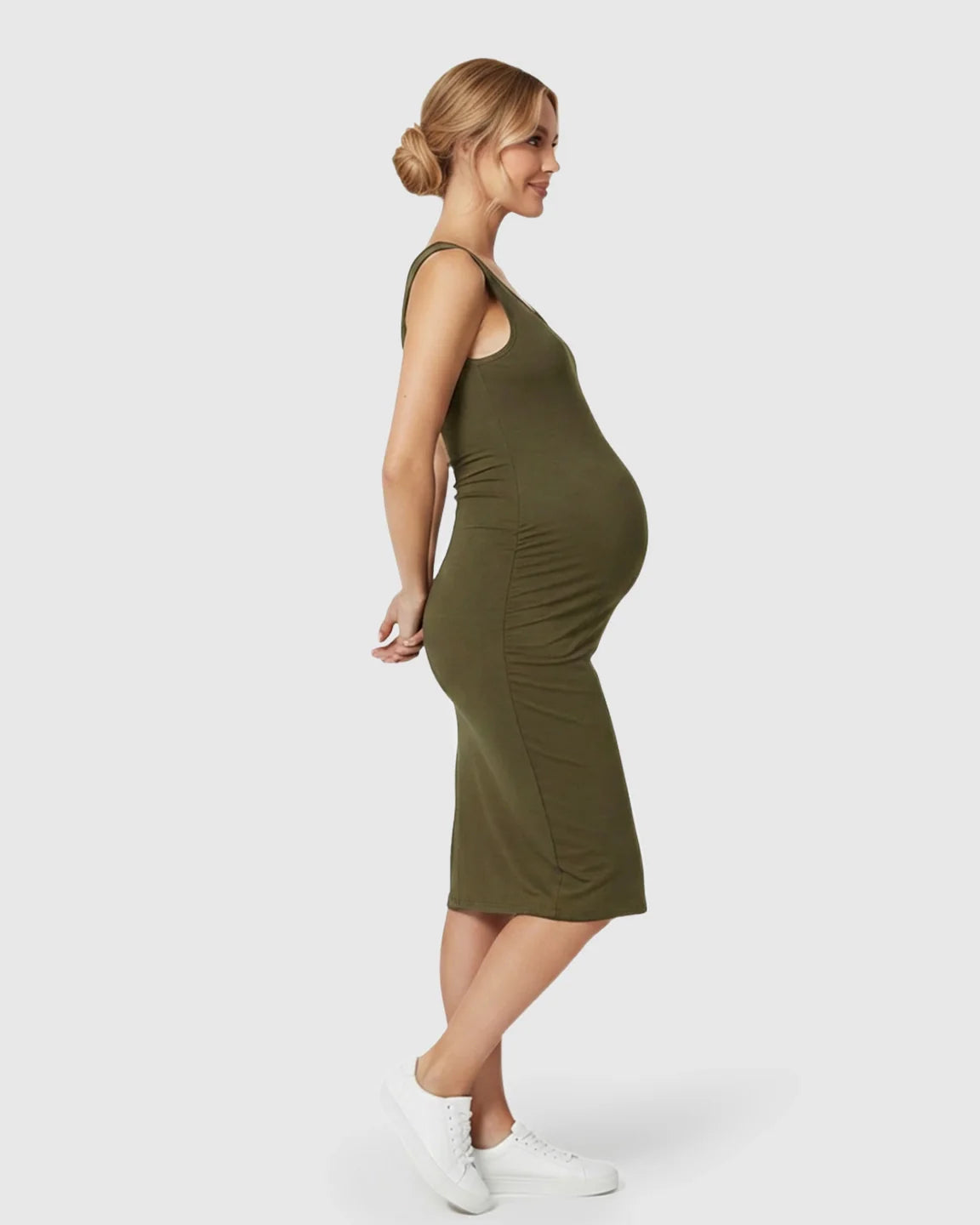Smiling pregnant woman in a sleeveless olive green bodycon maternity dress with side pleats and white sneakers.