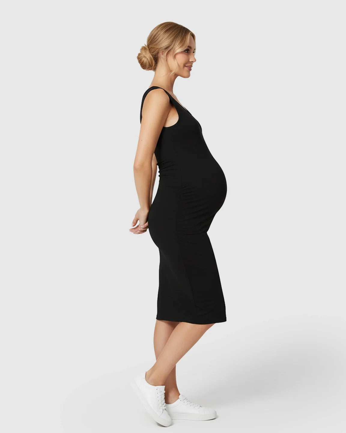 Smiling pregnant woman in a black sleeveless bodycon maternity dress with side pleats and white sneakers.