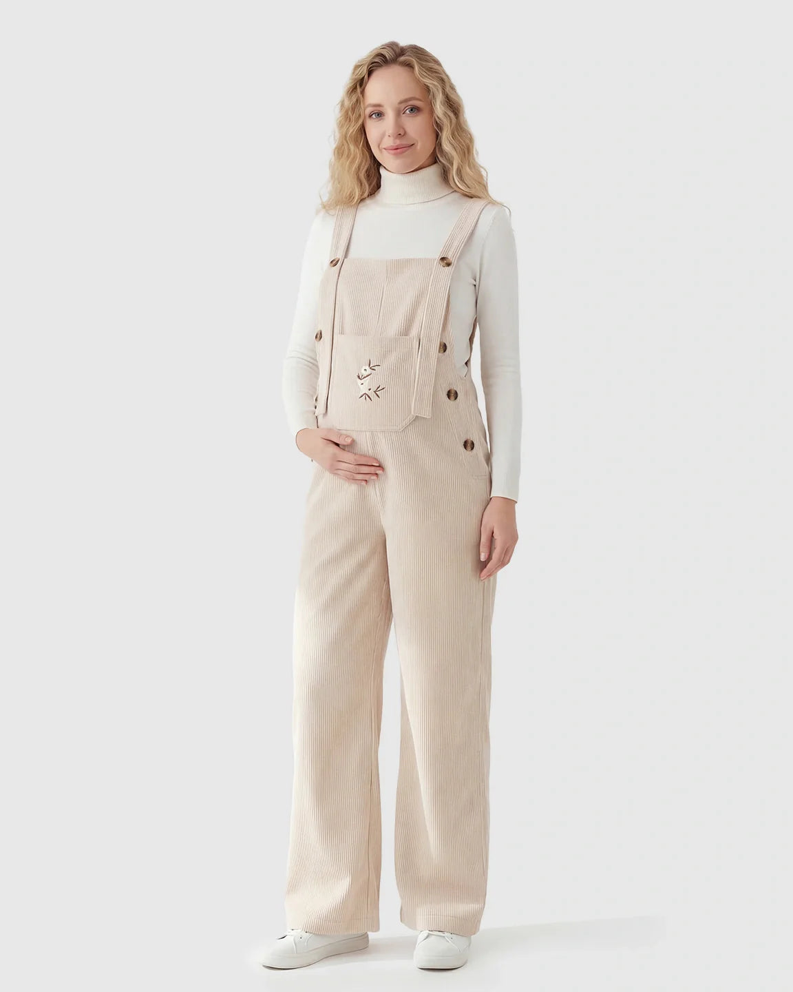 Blonde pregnant woman in beige corduroy maternity overalls with adjustable straps and cat embroidery.