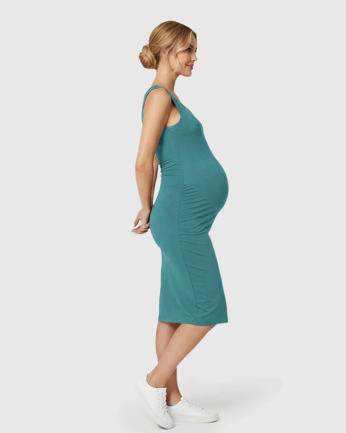 Smiling pregnant woman in a teal sleeveless bodycon maternity dress with side pleats and white sneakers.