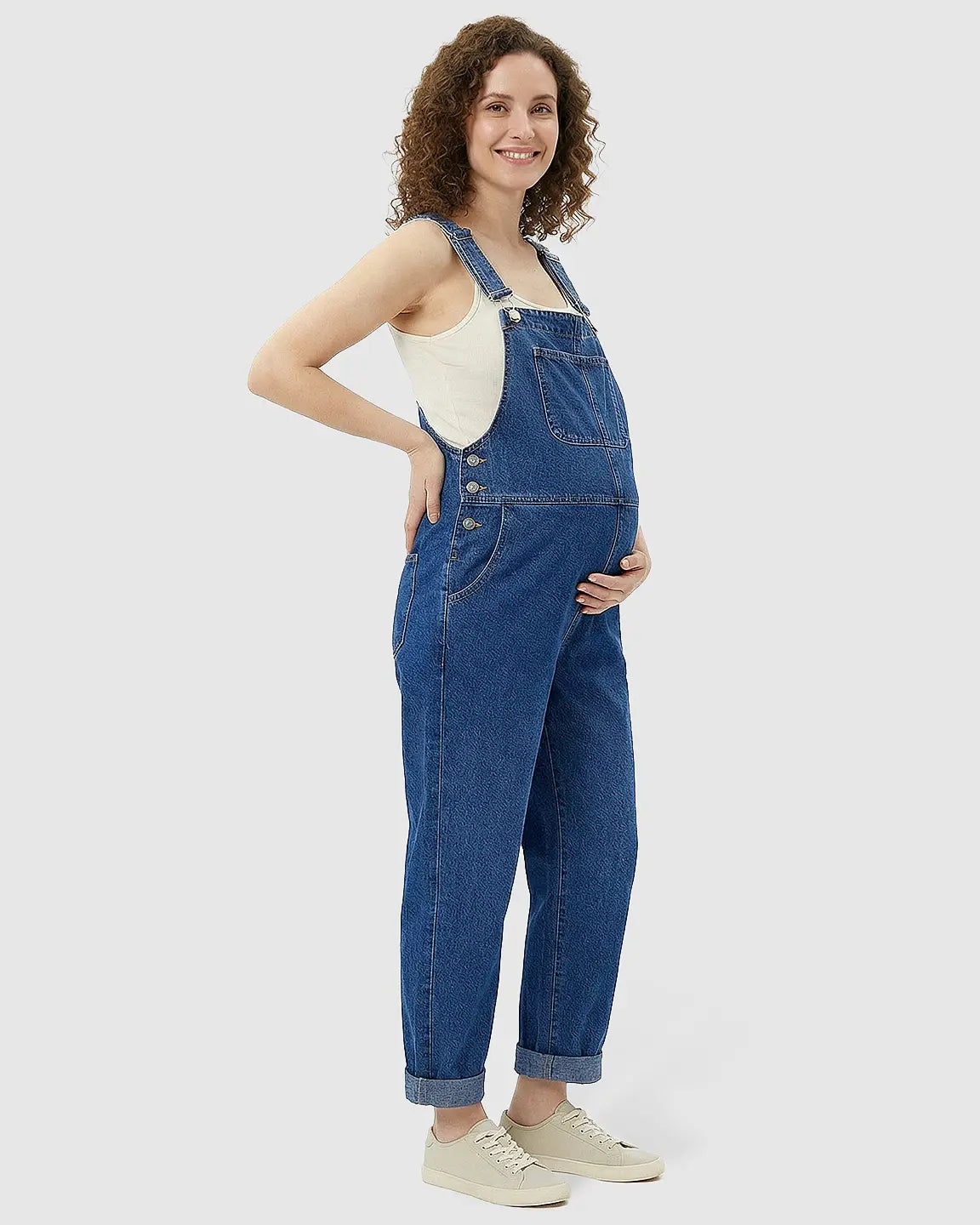 Smiling pregnant woman in a loose, blue denim maternity jumpsuit with adjustable straps and cuffed ankles.