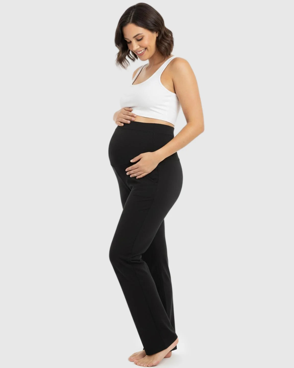 Smiling pregnant woman in black stretch flared maternity pants with over-belly waistband and a white crop top.