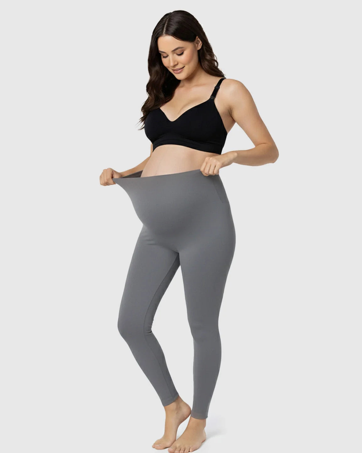 Smiling pregnant woman in a black bra pulling up gray stretch maternity leggings with a supportive belly panel.