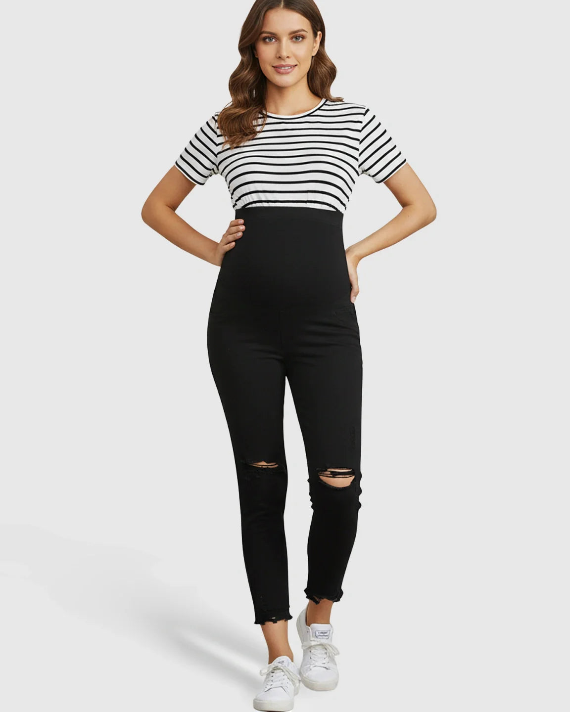 Maternity skinny jeans in black with distressed knees, ankle-fray hem, and over-belly support panel.