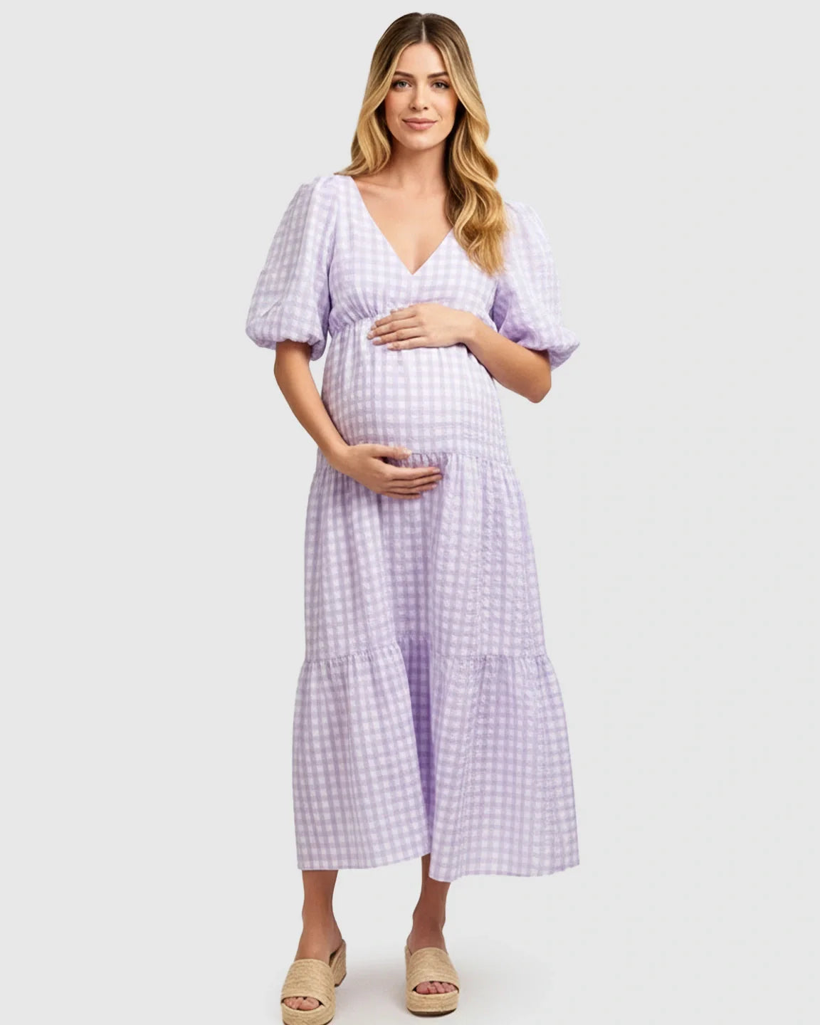 Pregnant woman in a lavender gingham puffed sleeve V-neck tiered maxi dress, holding her bump.