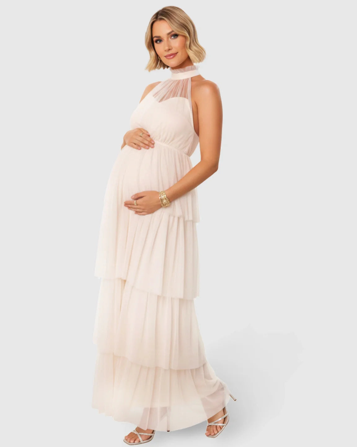 Pregnant woman in a blush pink halter-neck tiered mesh maternity gown, holding her bump.