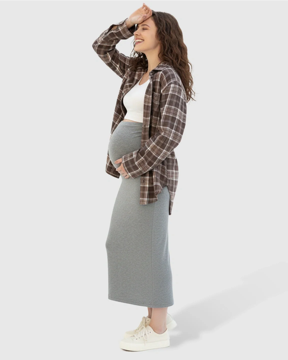 Smiling pregnant woman in a gray midi skirt, white crop top, and brown plaid shirt.