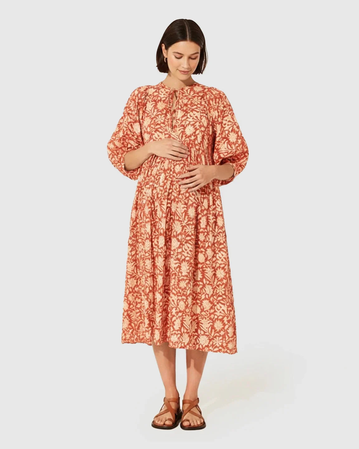 Pregnant woman in a loose-fit terracotta floral maternity midi dress with puff sleeves and knot detail.