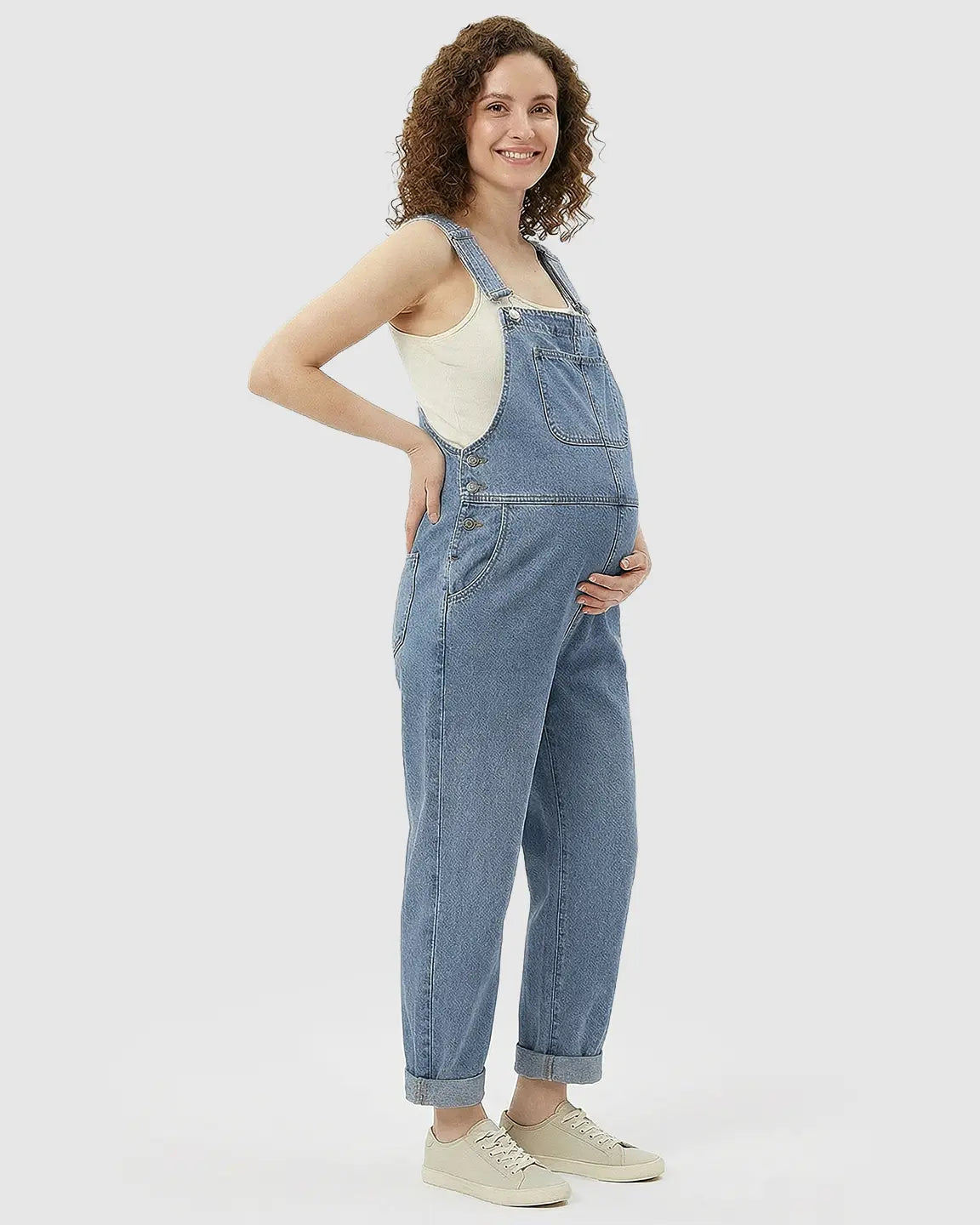 Smiling pregnant woman in a loose-fit, light blue denim maternity jumpsuit with adjustable straps and cuffed ankles.