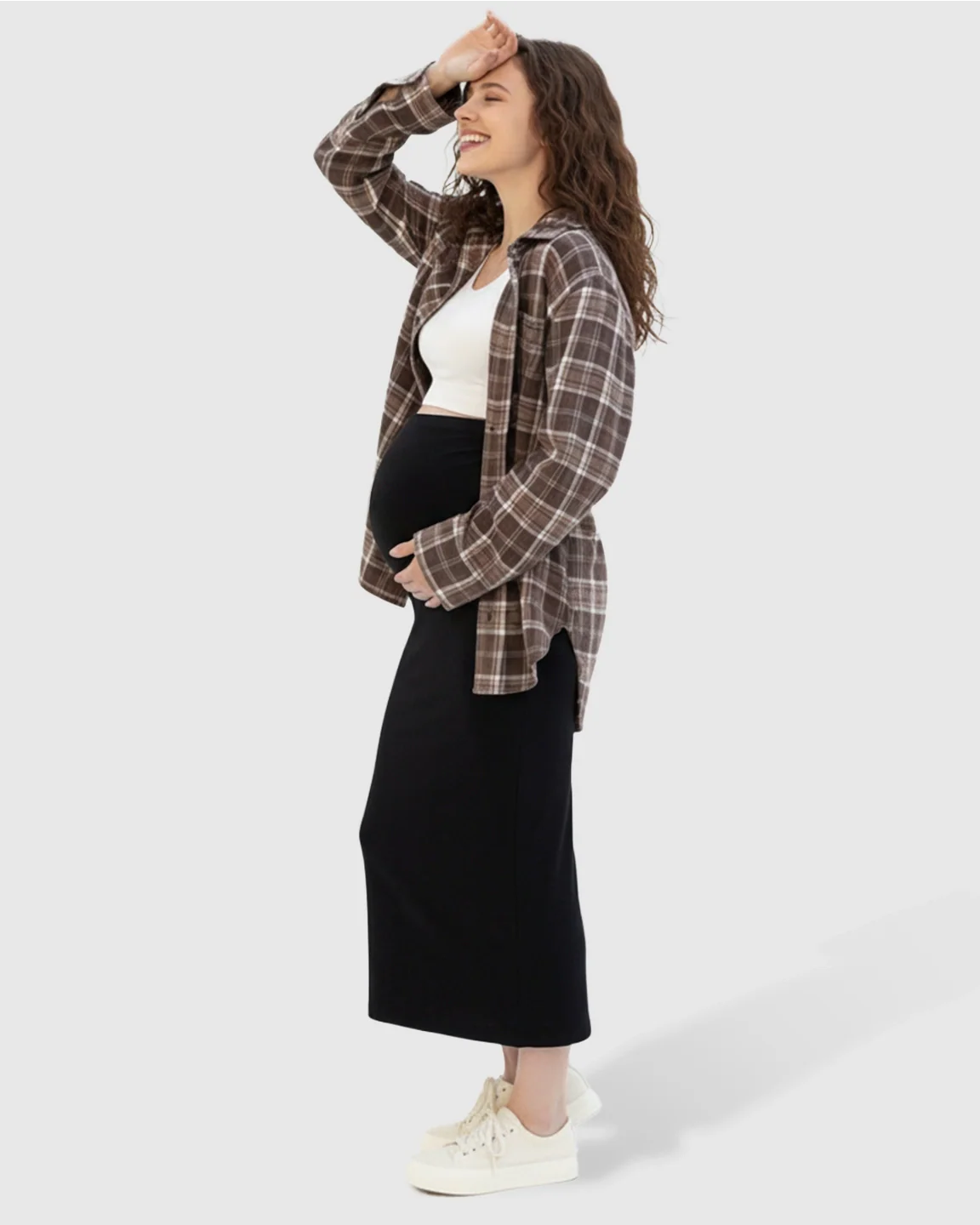 Smiling pregnant woman in a black midi skirt, white crop top, and brown plaid shirt.
