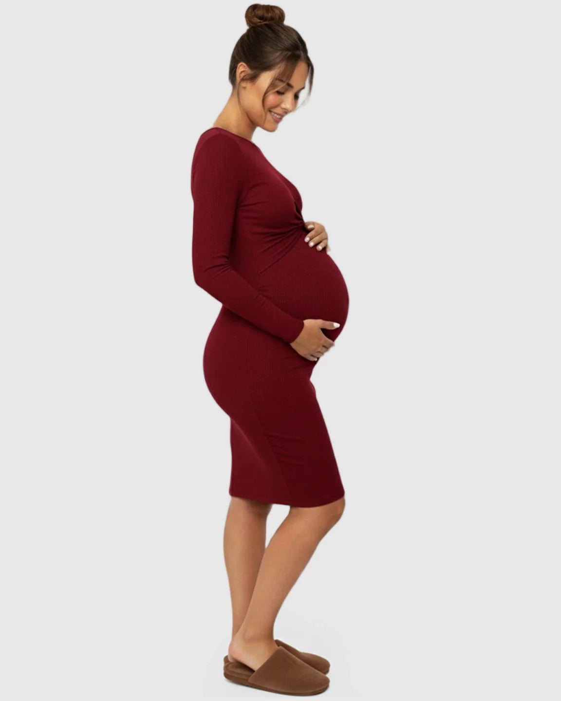 Pregnant woman in a red ribbed bodycon maternity dress with a twist-front detail, smiling and holding her bump.