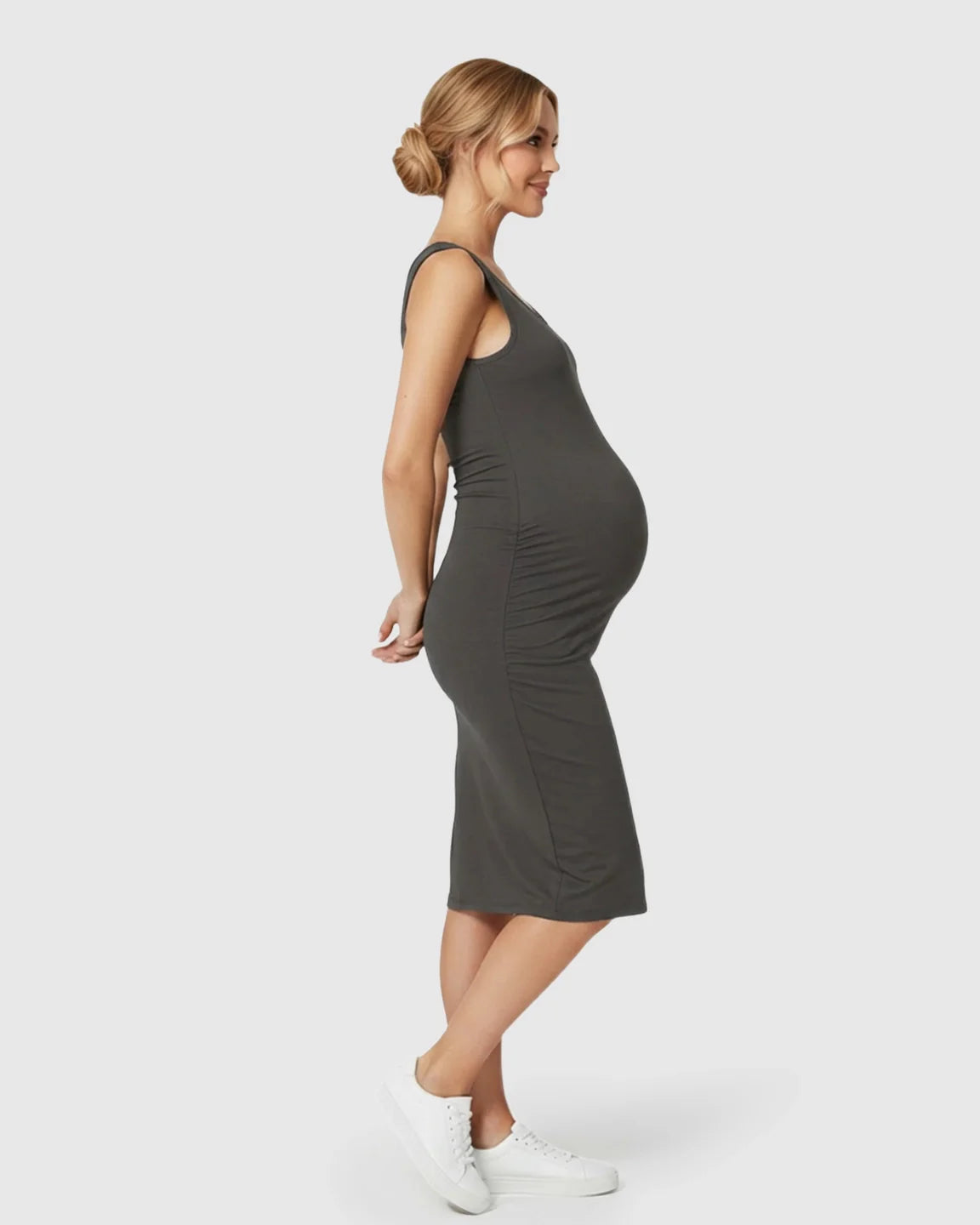 Smiling pregnant woman in a charcoal sleeveless bodycon maternity dress with side pleats and white sneakers.