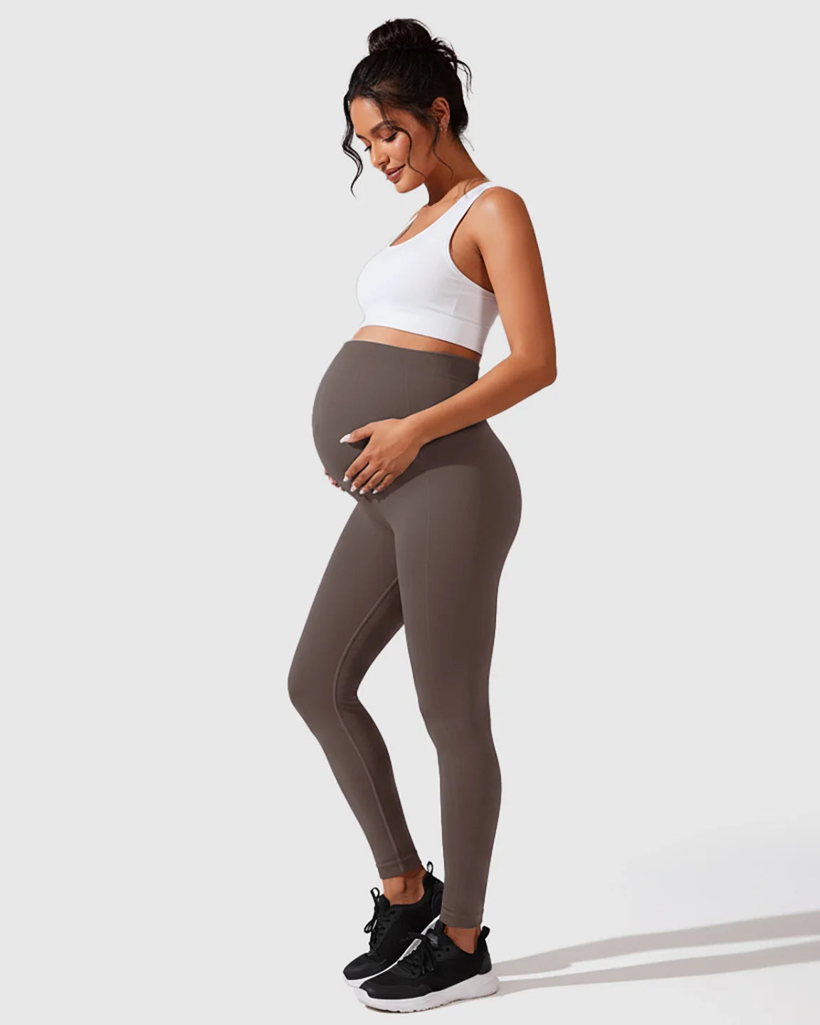 Pregnant woman in seamless, high-waisted gray maternity leggings and a white crop top, cradling her bump.