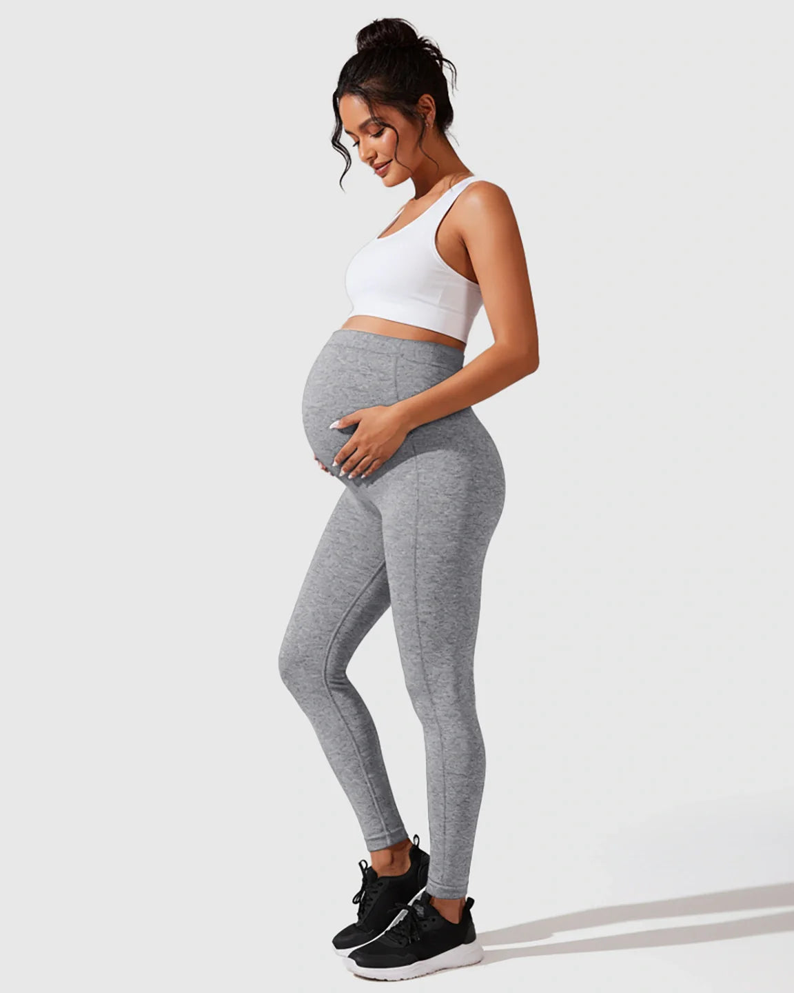 Pregnant woman in gray seamless M-shape maternity leggings and a white crop top, cradling her bump.