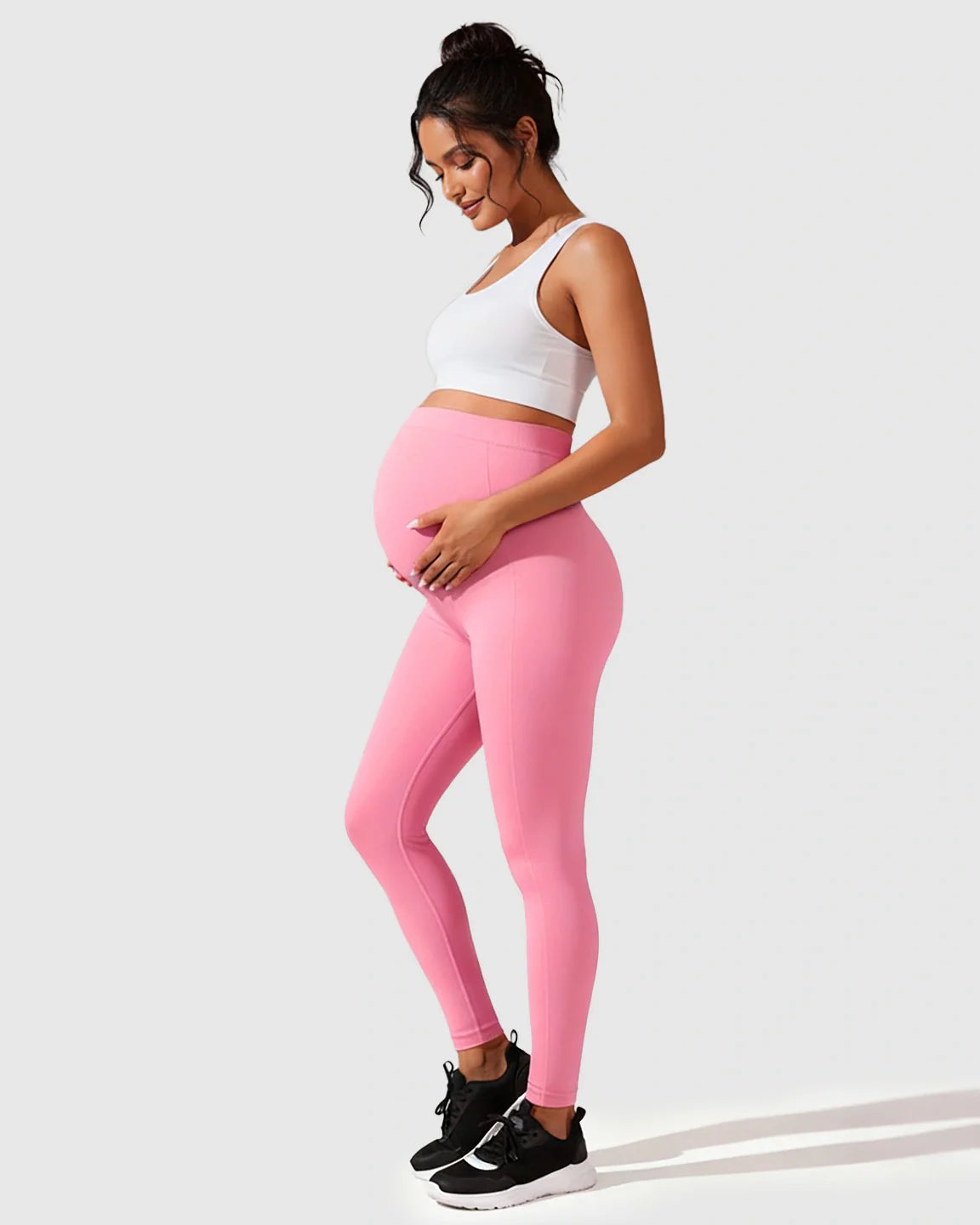 Pregnant woman in pink seamless M-shape maternity leggings and a white crop top, cradling her bump.