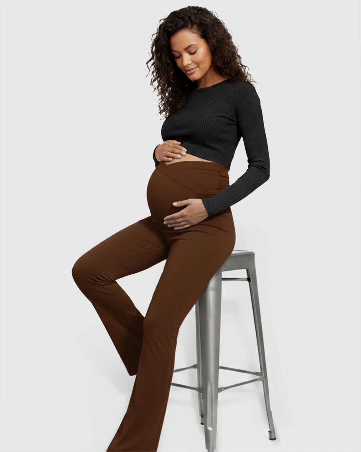 Pregnant woman in brown crossed waistband flared maternity pants and a black ribbed crop top.