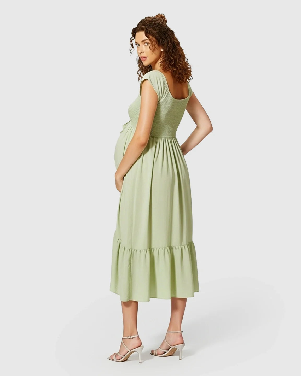 Pregnant woman in a sage green smocked-bodice flowy maternity midi dress with cap sleeves and a tiered skirt.
