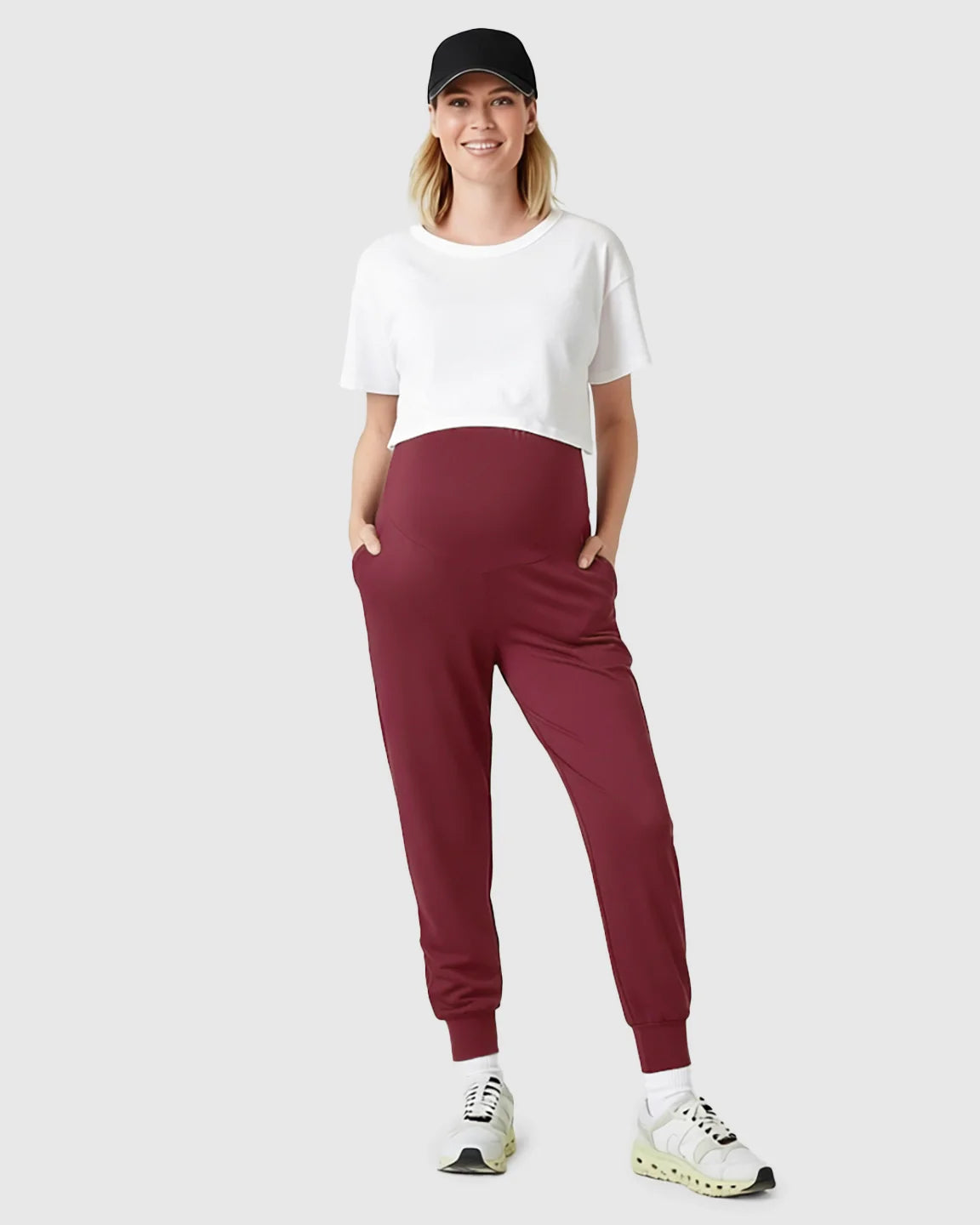 Woman in black cap, white crop top, and supportive slim-fitting burgundy maternity joggers with pockets.