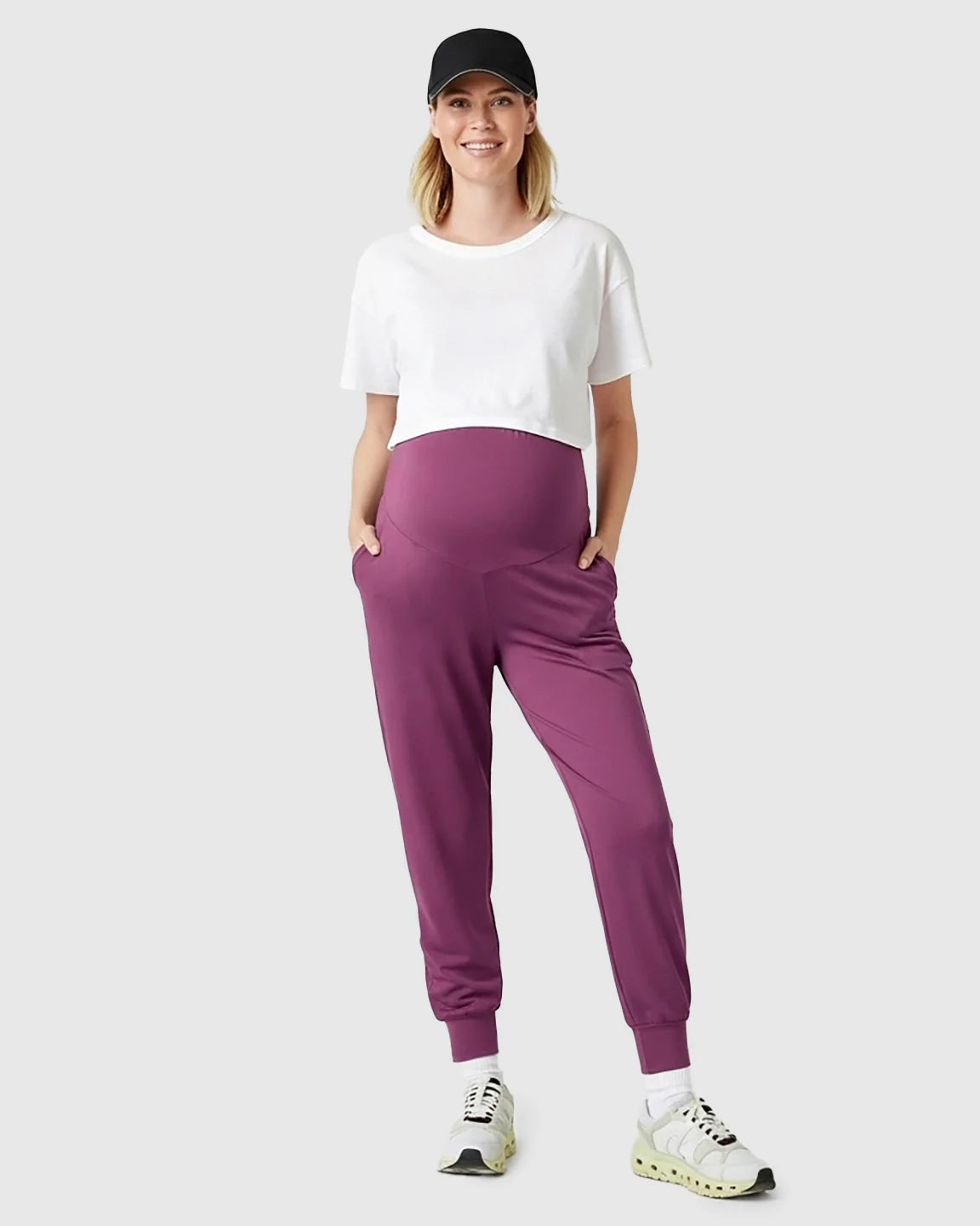 Smiling pregnant woman in plum supportive maternity joggers, white crop top, and black cap.