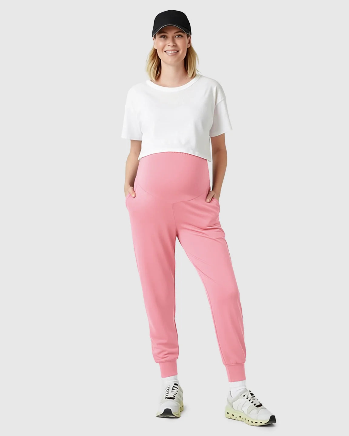 Smiling pregnant woman in pink supportive maternity joggers, white crop top, and black cap.