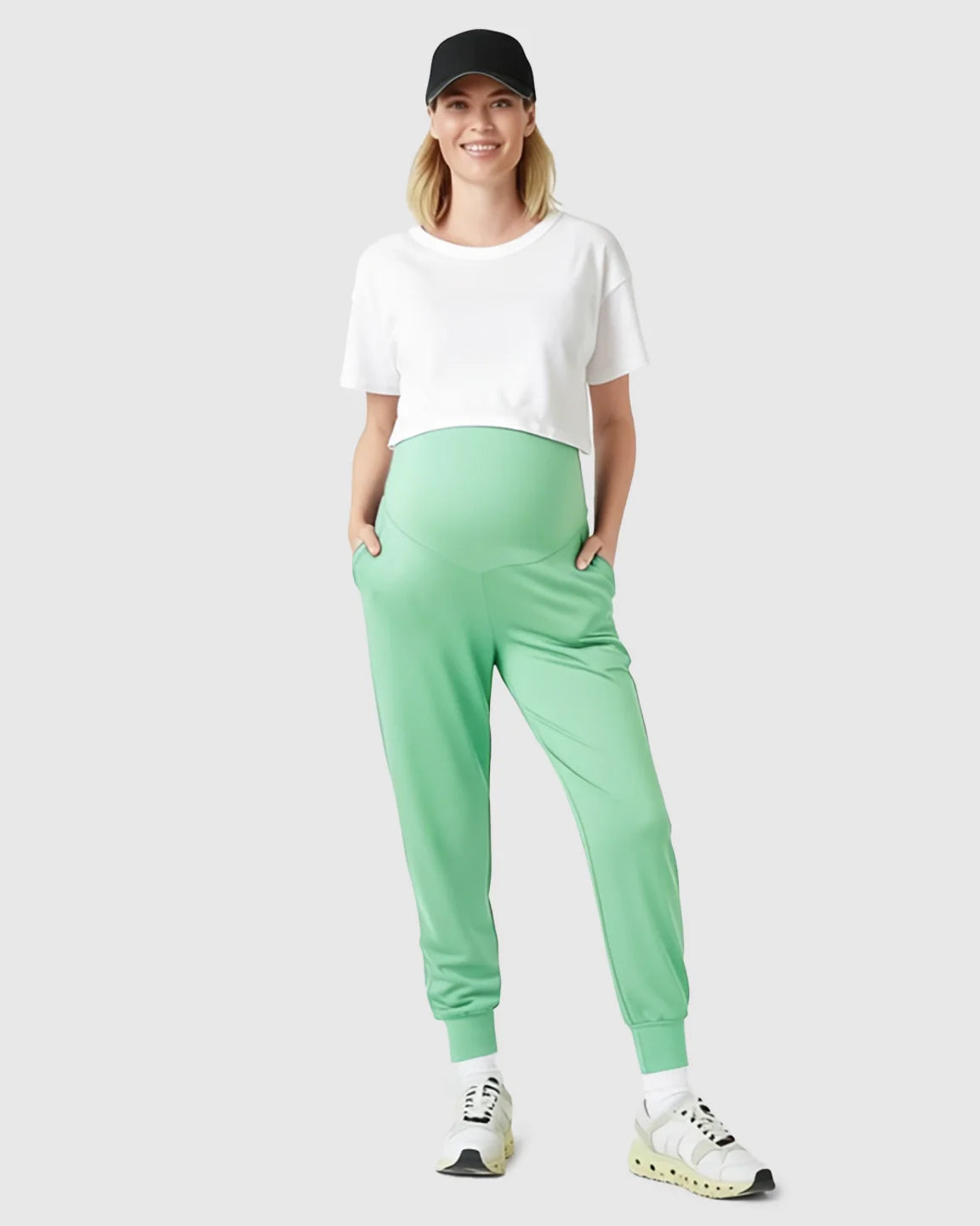 Smiling pregnant woman in mint green supportive maternity joggers, white crop top, and black cap.