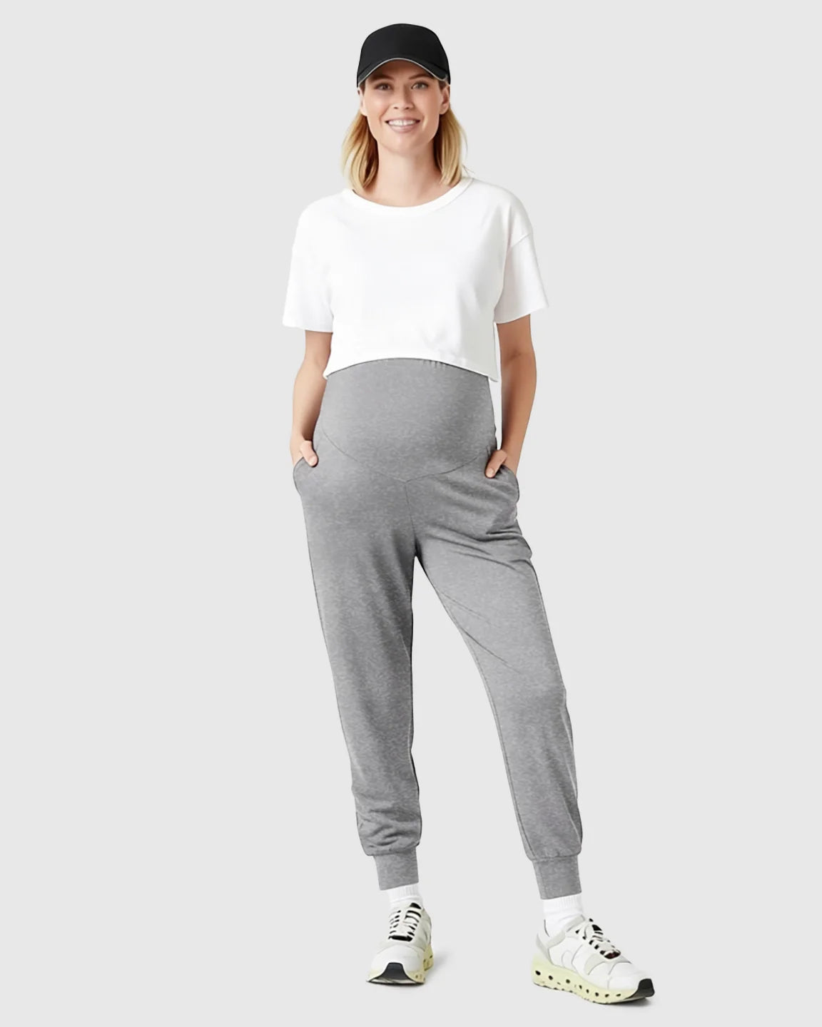 Smiling pregnant woman in gray supportive slim-fitting maternity joggers, white crop top, and black cap.