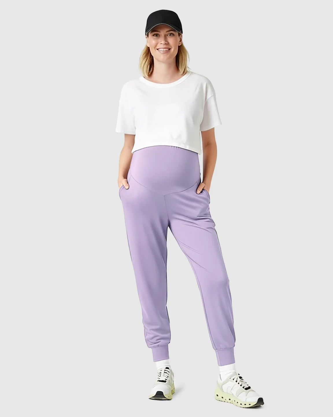 Smiling pregnant woman in supportive lavender maternity joggers, white crop top, and black baseball cap.