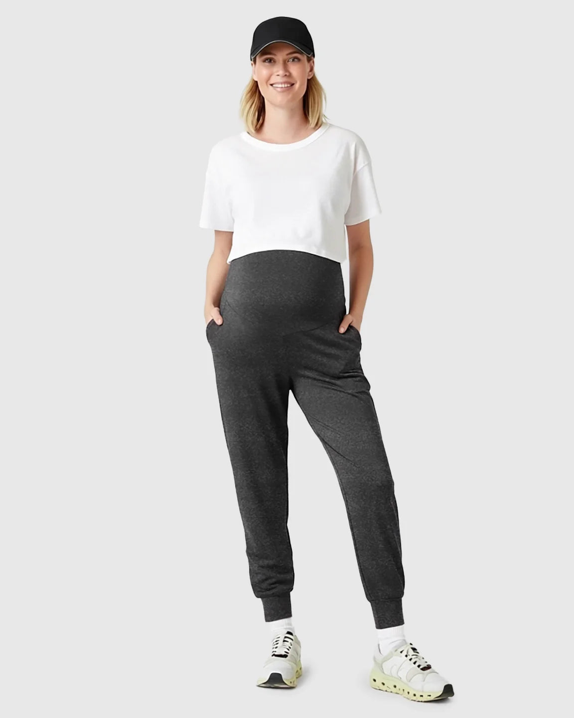 Smiling pregnant woman in black cap, white tee, and charcoal supportive slim-fitting maternity joggers.