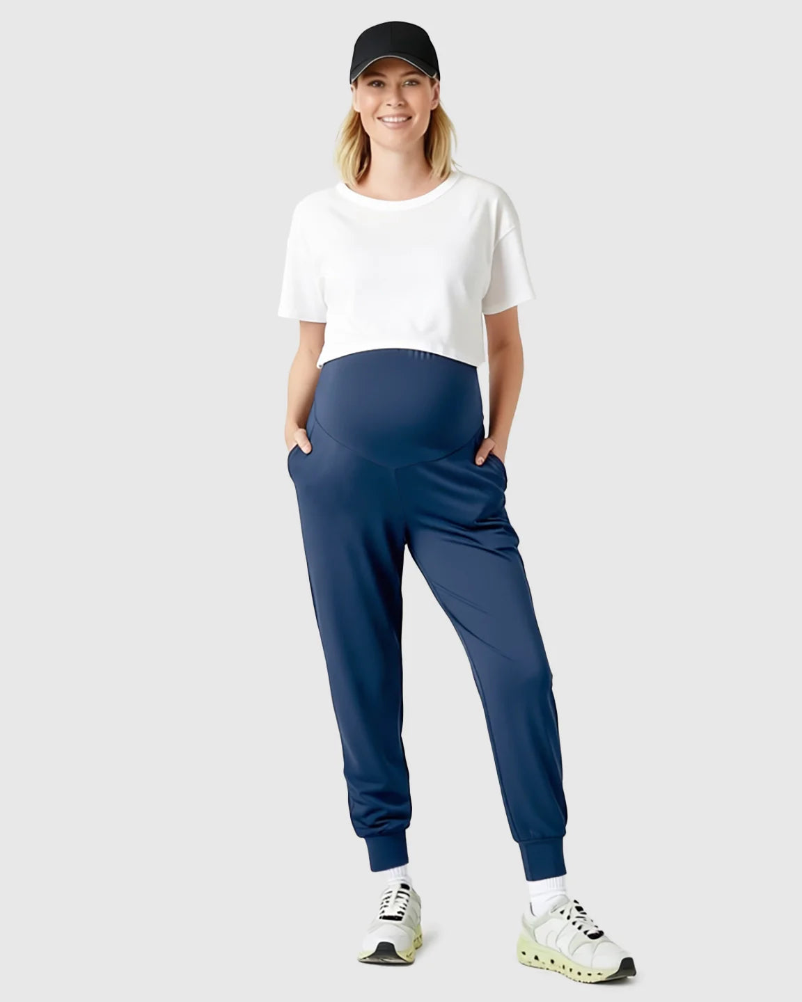 Smiling pregnant woman in navy supportive slim-fitting maternity joggers, white crop top, and black cap.