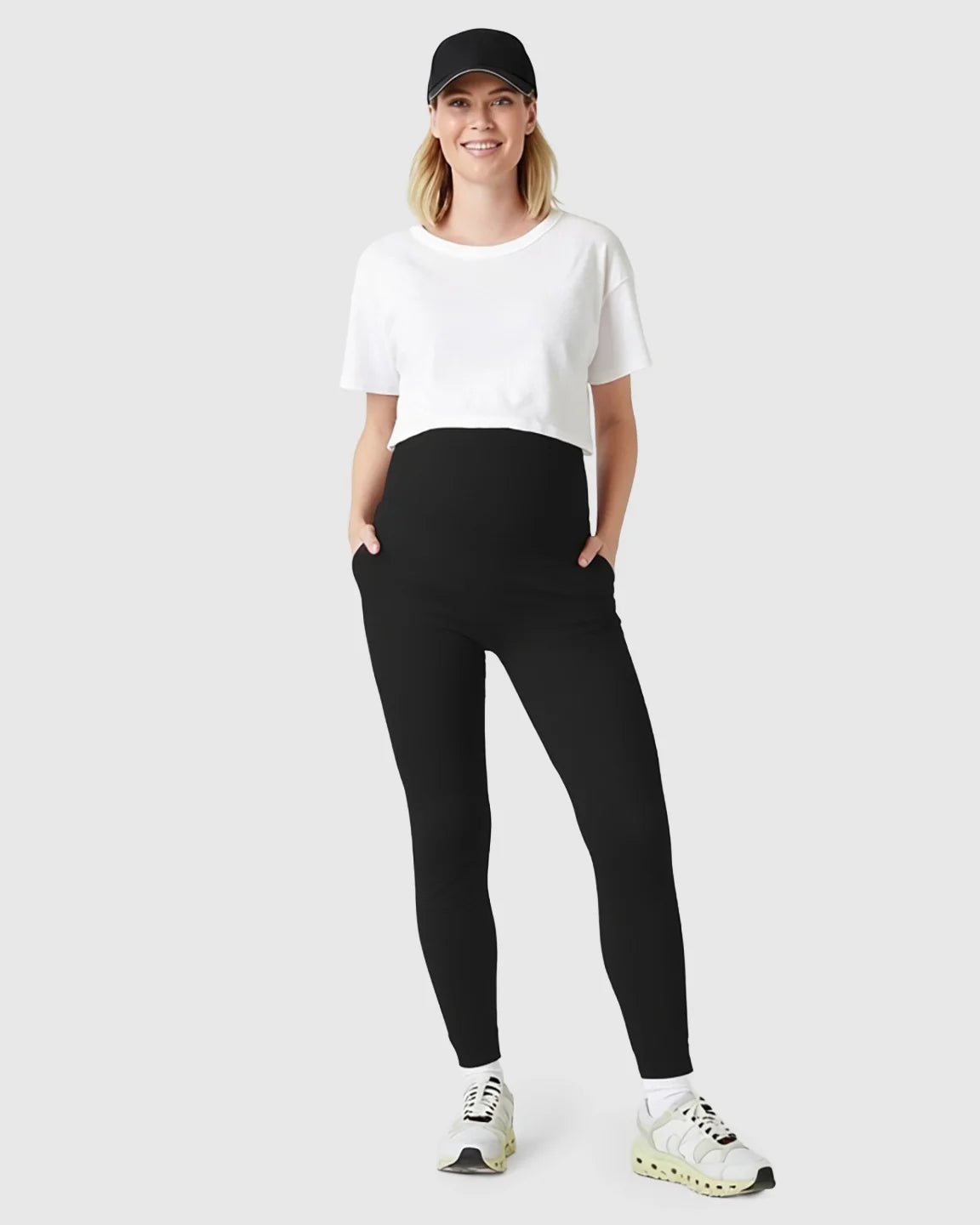 Smiling pregnant woman in black supportive slim-fitting maternity joggers, white crop top, and black cap.