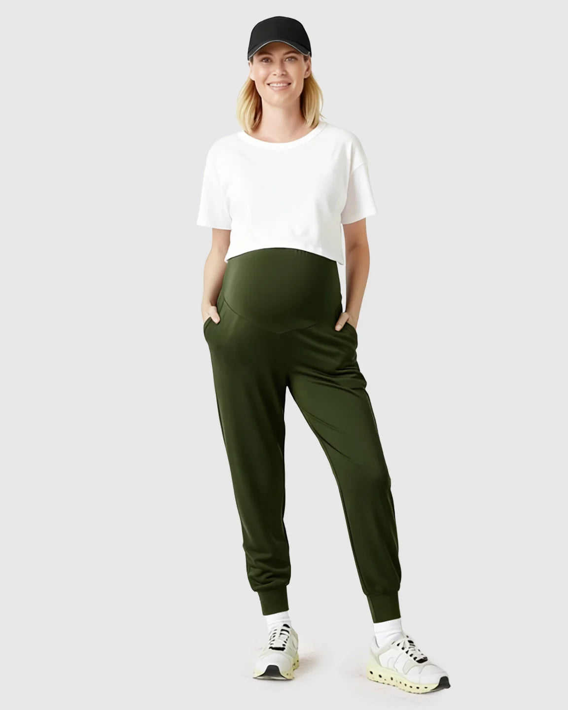 Smiling pregnant woman in olive green supportive maternity joggers, white crop top, and black cap.
