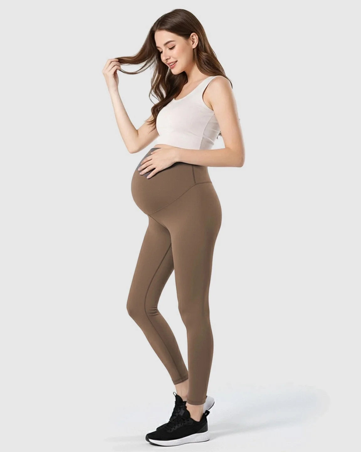 Smiling pregnant woman in brown double-layer belly support maternity leggings and white tank top.