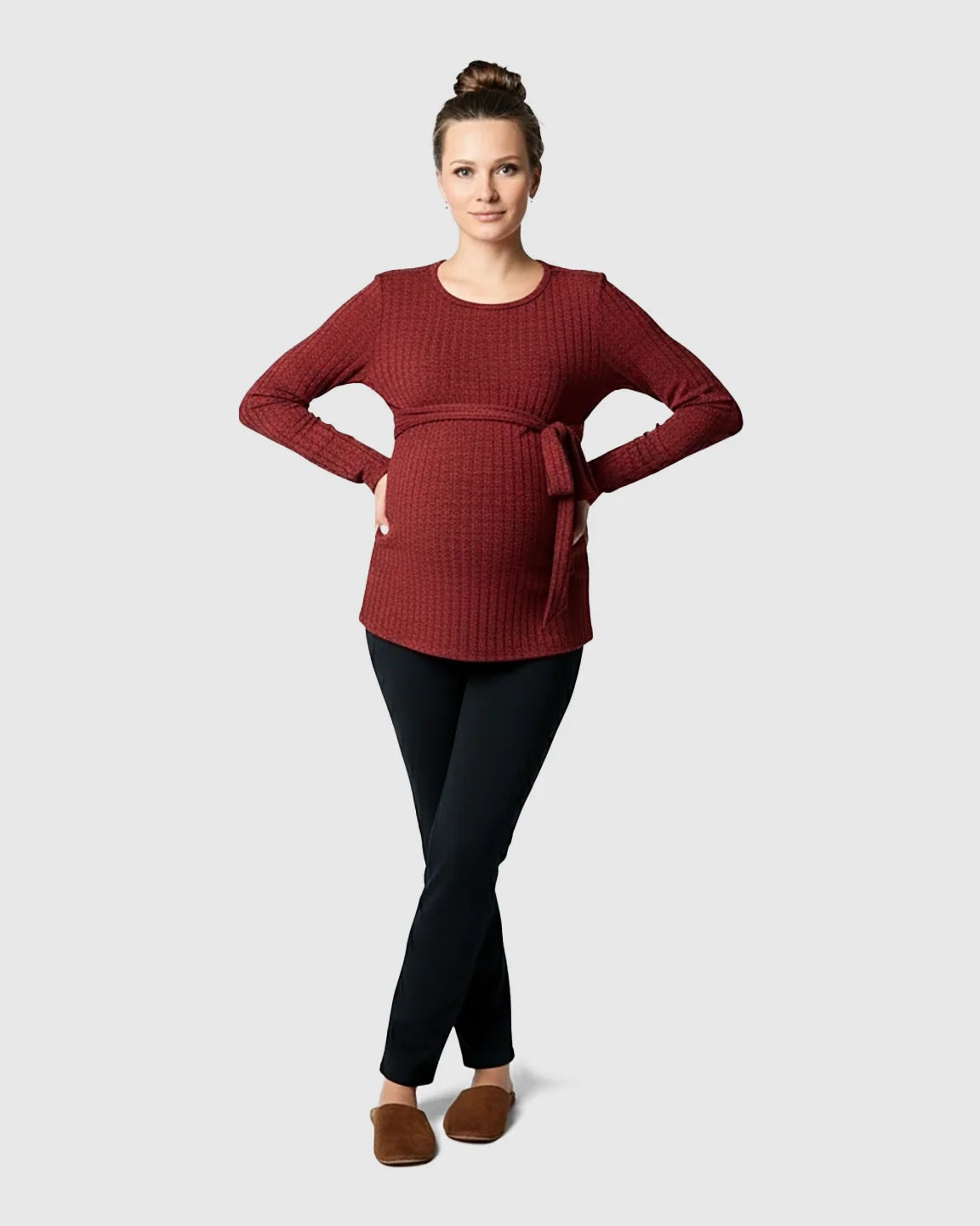Woman in a ribbed, rust-colored maternity sweater with a tie-waist, black pants, and brown slippers.