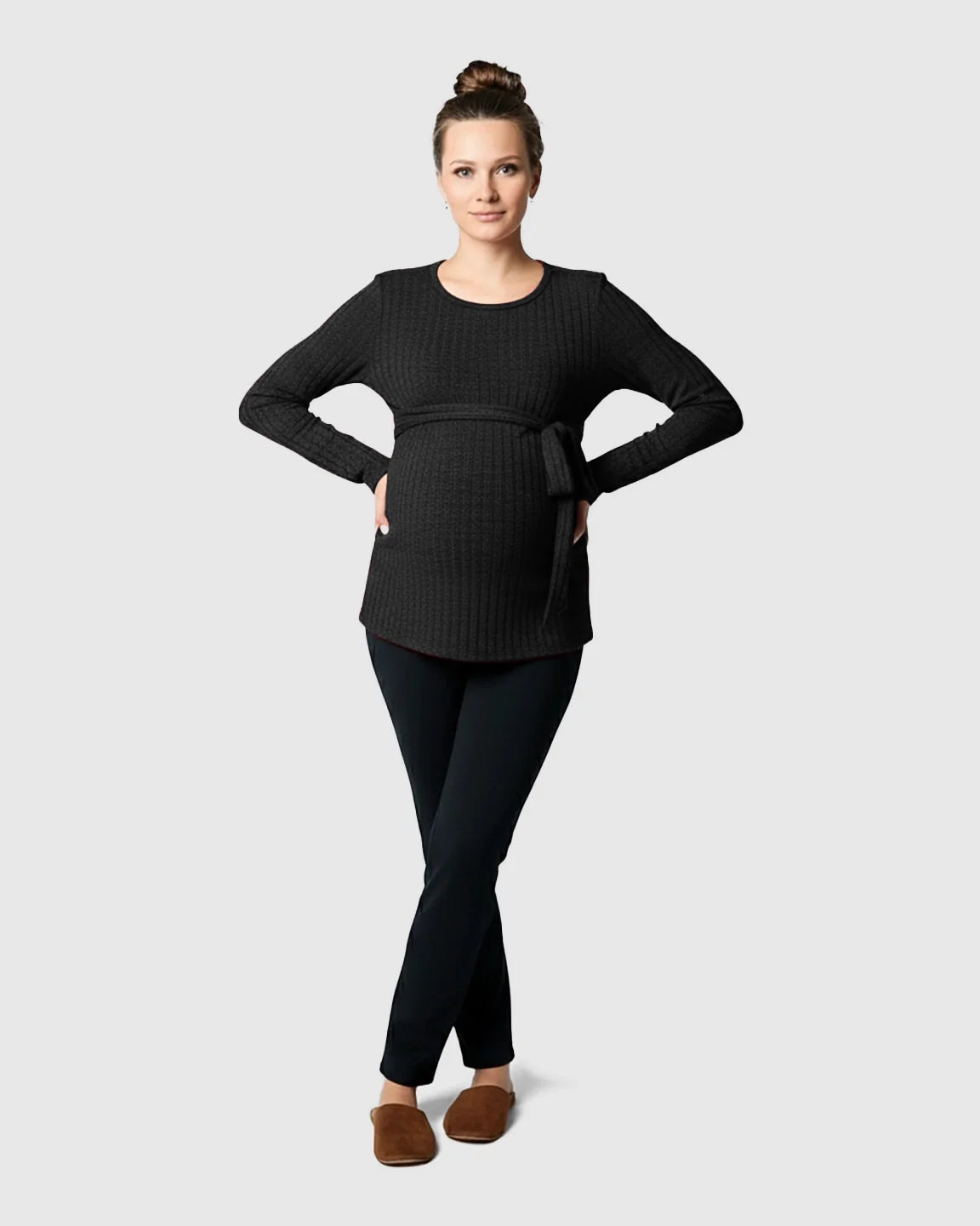 Woman in a dark gray ribbed maternity sweater with a tie-waist, black pants, and brown slippers.