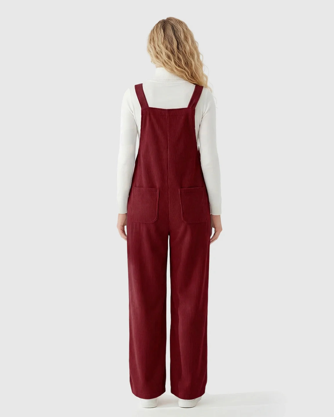 Woman from behind in red corduroy maternity overalls with adjustable straps and front pockets over a white turtleneck.