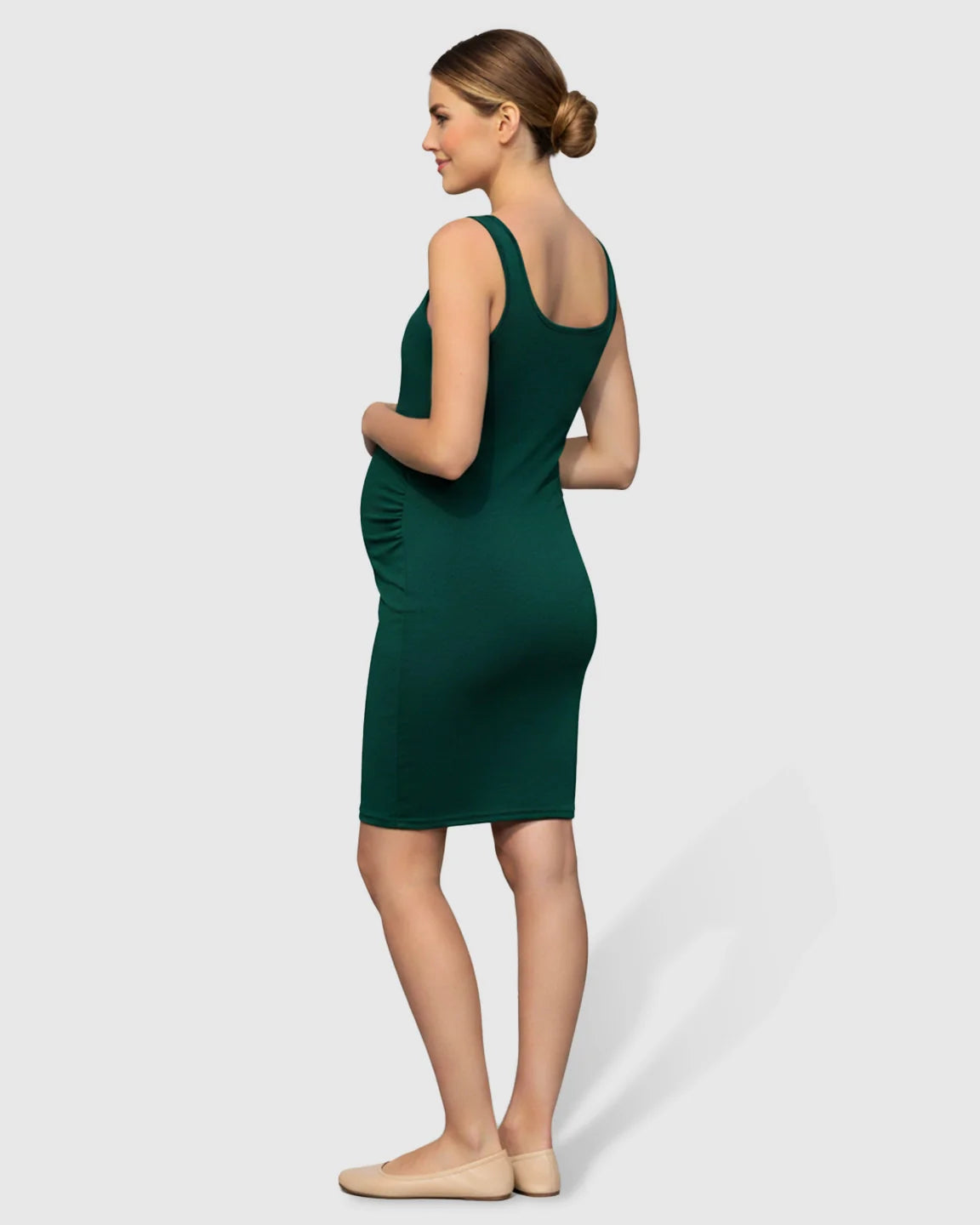 Pregnant woman in a dark green, scoop neck, side-ruched bodycon maternity dress with nude flats.