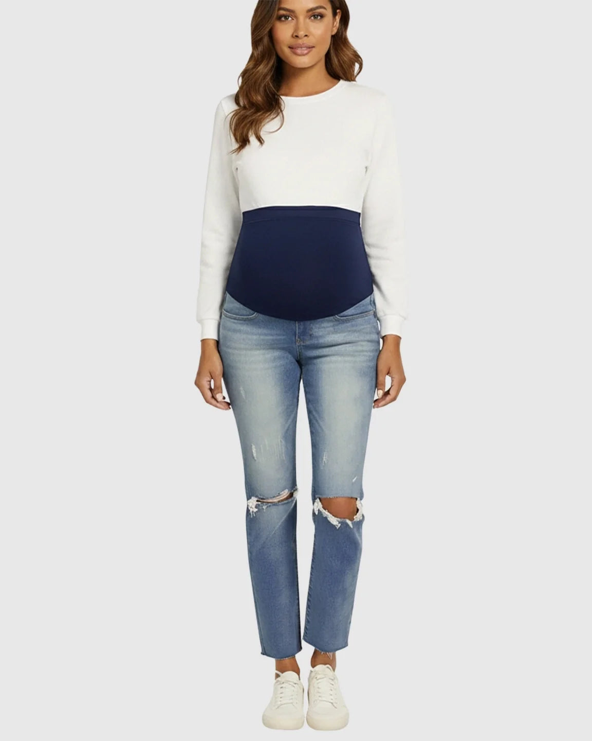 Woman wearing light wash distressed maternity jeans with a navy belly band and white long-sleeve top.