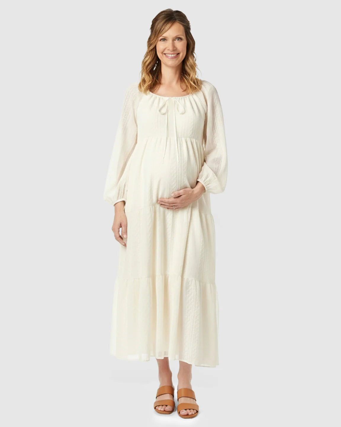 Smiling pregnant woman in a cream textured long-sleeve tiered maternity midi dress with a tie-front neckline.