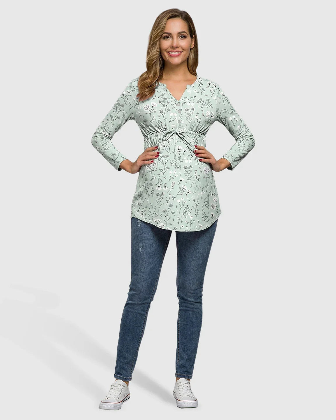 Smiling pregnant woman in a light green floral notch neck maternity tunic with a drawstring waist and blue jeans.