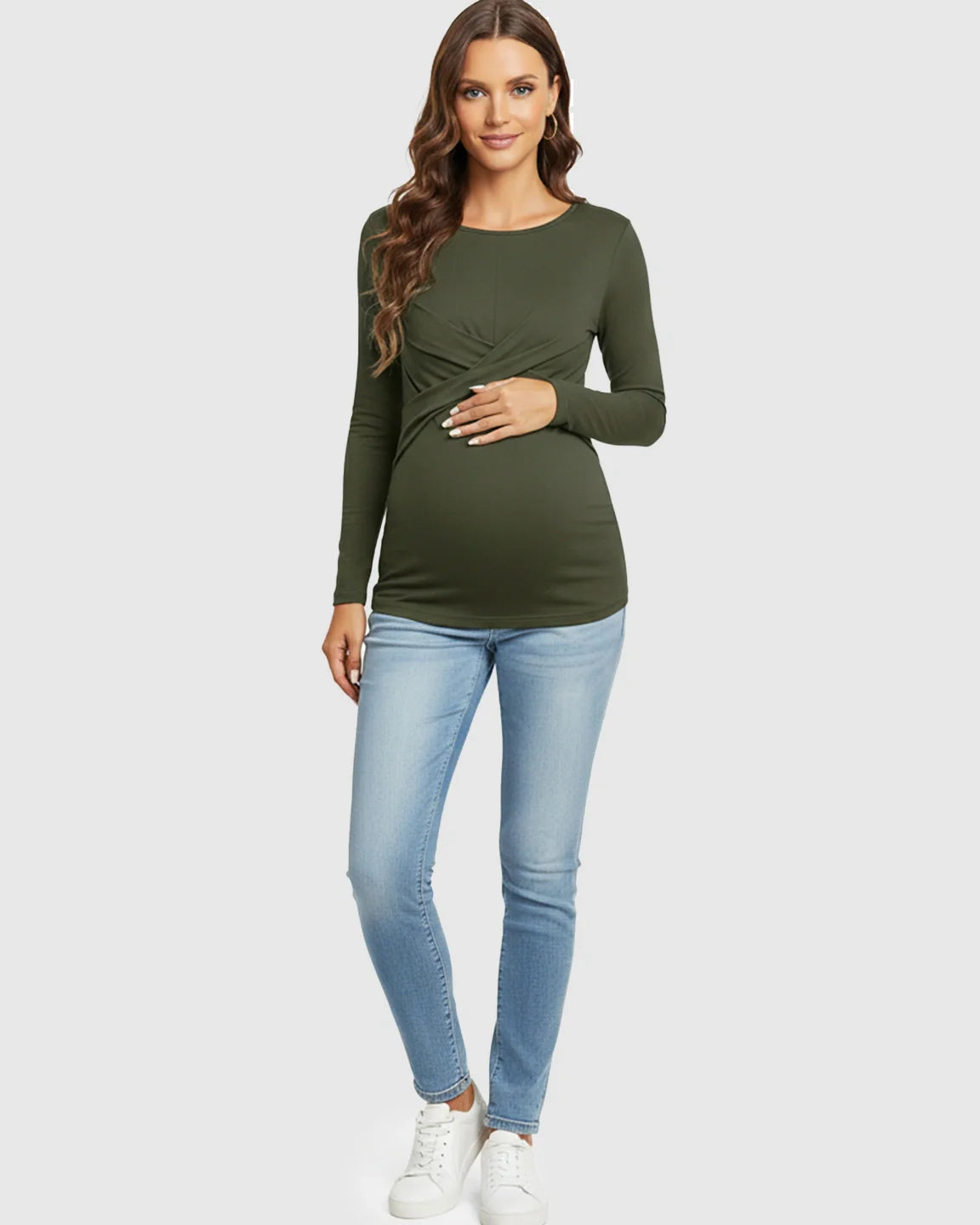 Maternity model in a dark olive green cross-pleat fitted long sleeve top, light wash jeans, and white sneakers.