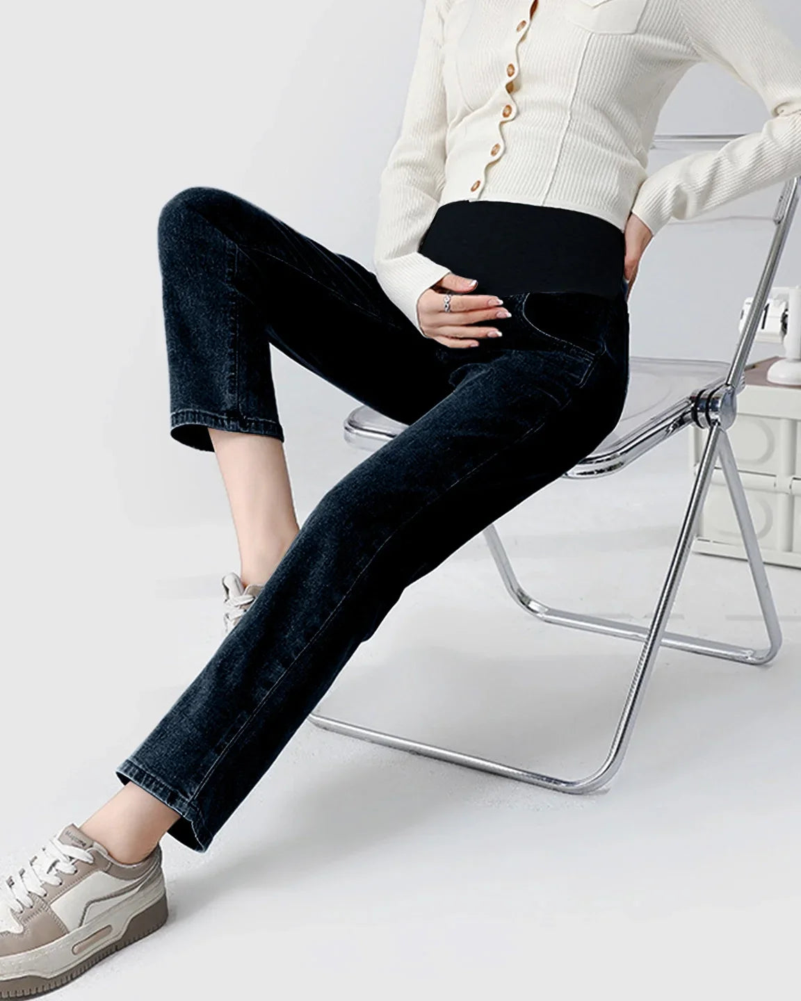 Woman wearing dark wash straight-leg maternity jeans with a black over-the-belly panel, sitting on a clear chair.