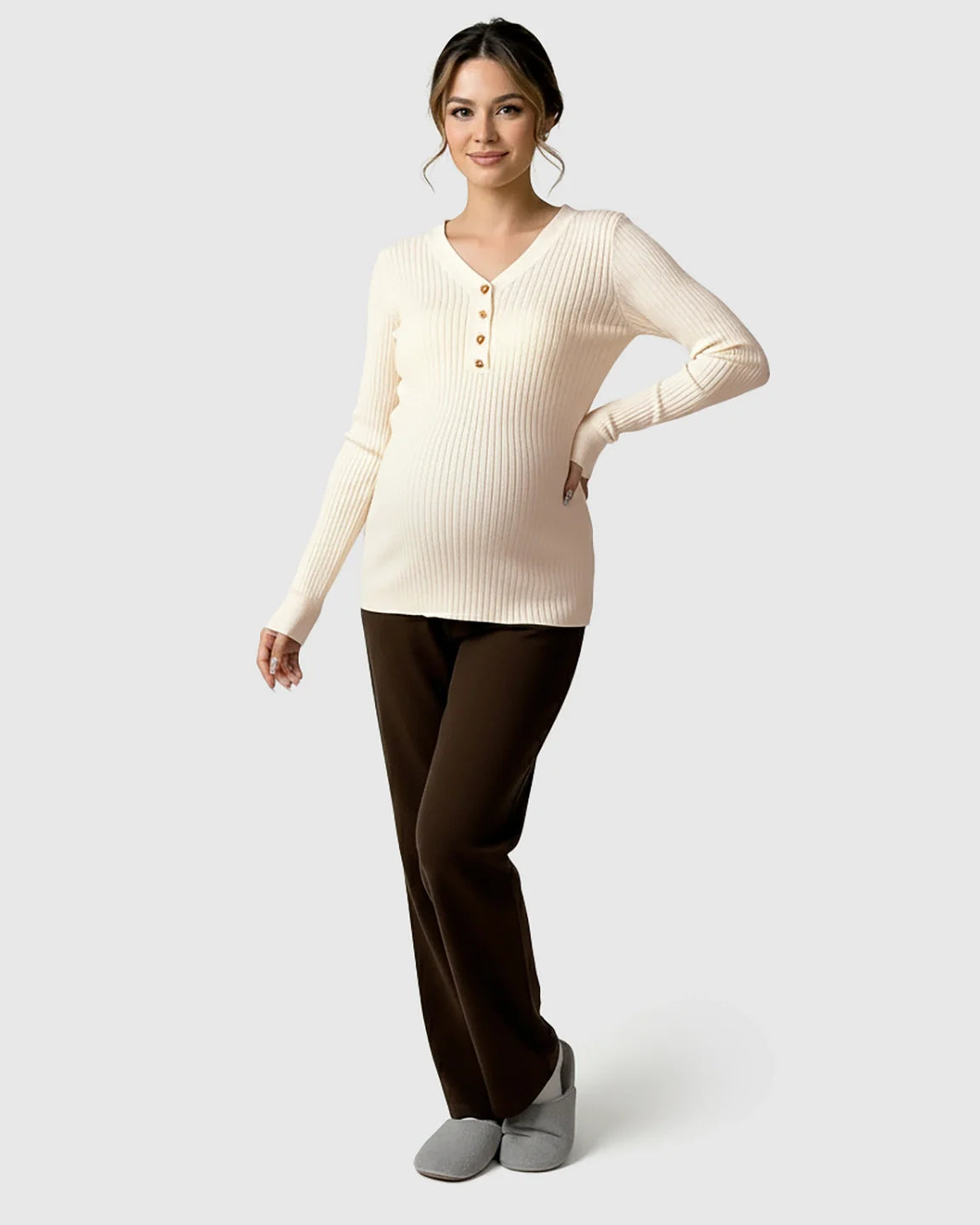 Pregnant woman in a cream ribbed V-neck maternity nursing sweater with gold buttons and brown pants.