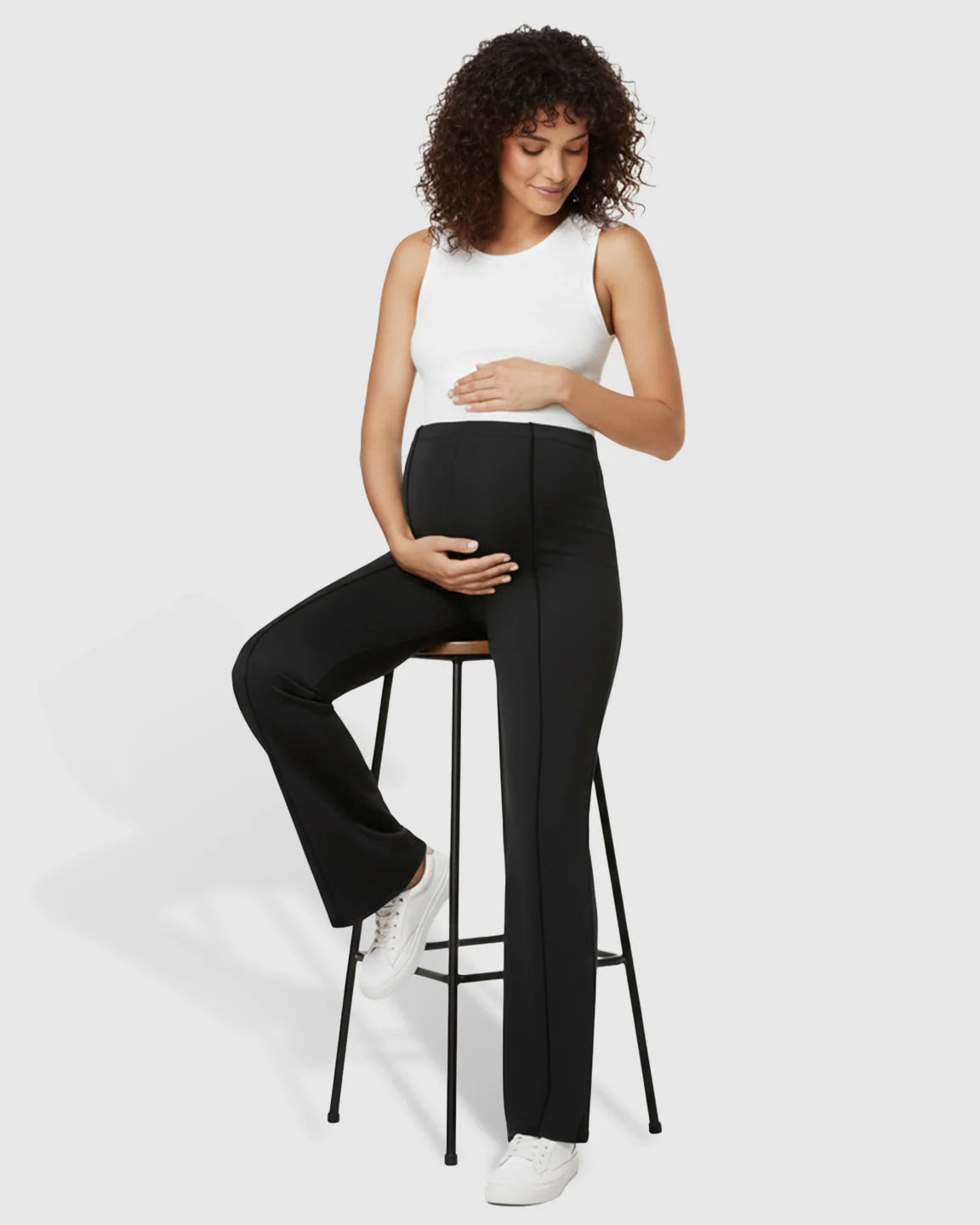 Pregnant woman in black butt-lifting flared maternity pants with white tank top, sitting on a stool.