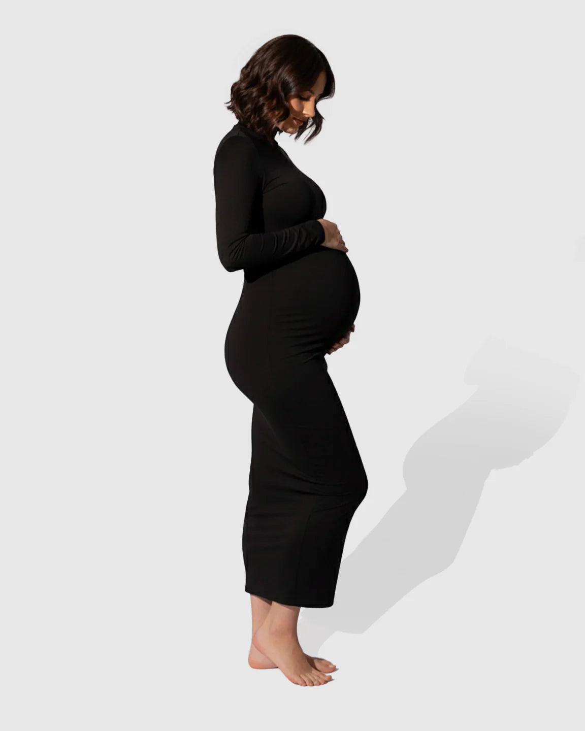 Woman in a black mock-neck bodycon maternity maxi dress, gently cradling her bump.