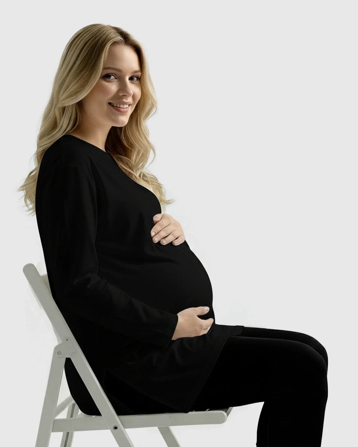 Smiling pregnant woman in a black long-sleeve soft cotton maternity top and black leggings, sitting on a white chair.