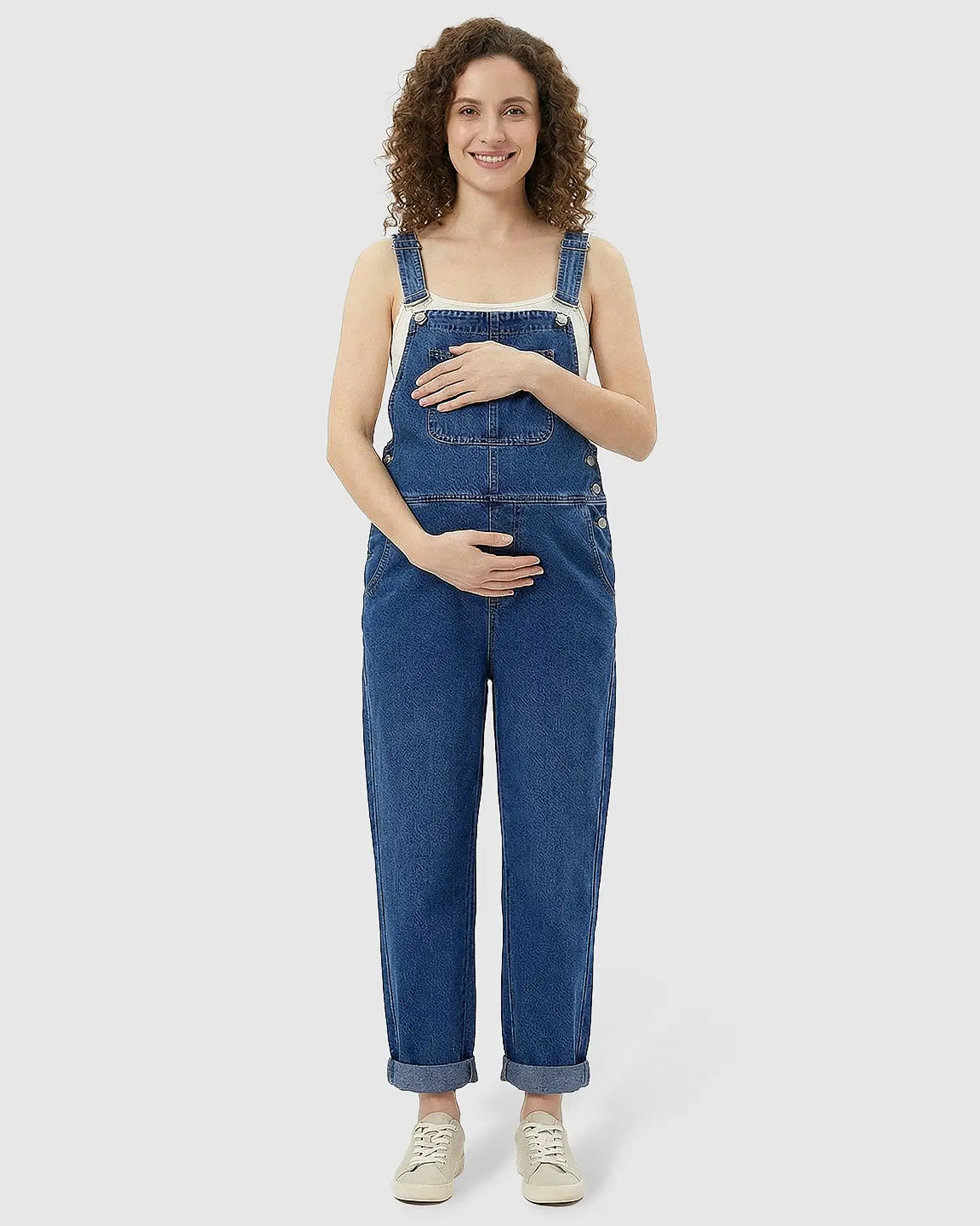 Smiling pregnant woman in a blue denim maternity jumpsuit with adjustable straps, cuffed ankles, and a white tank top.
