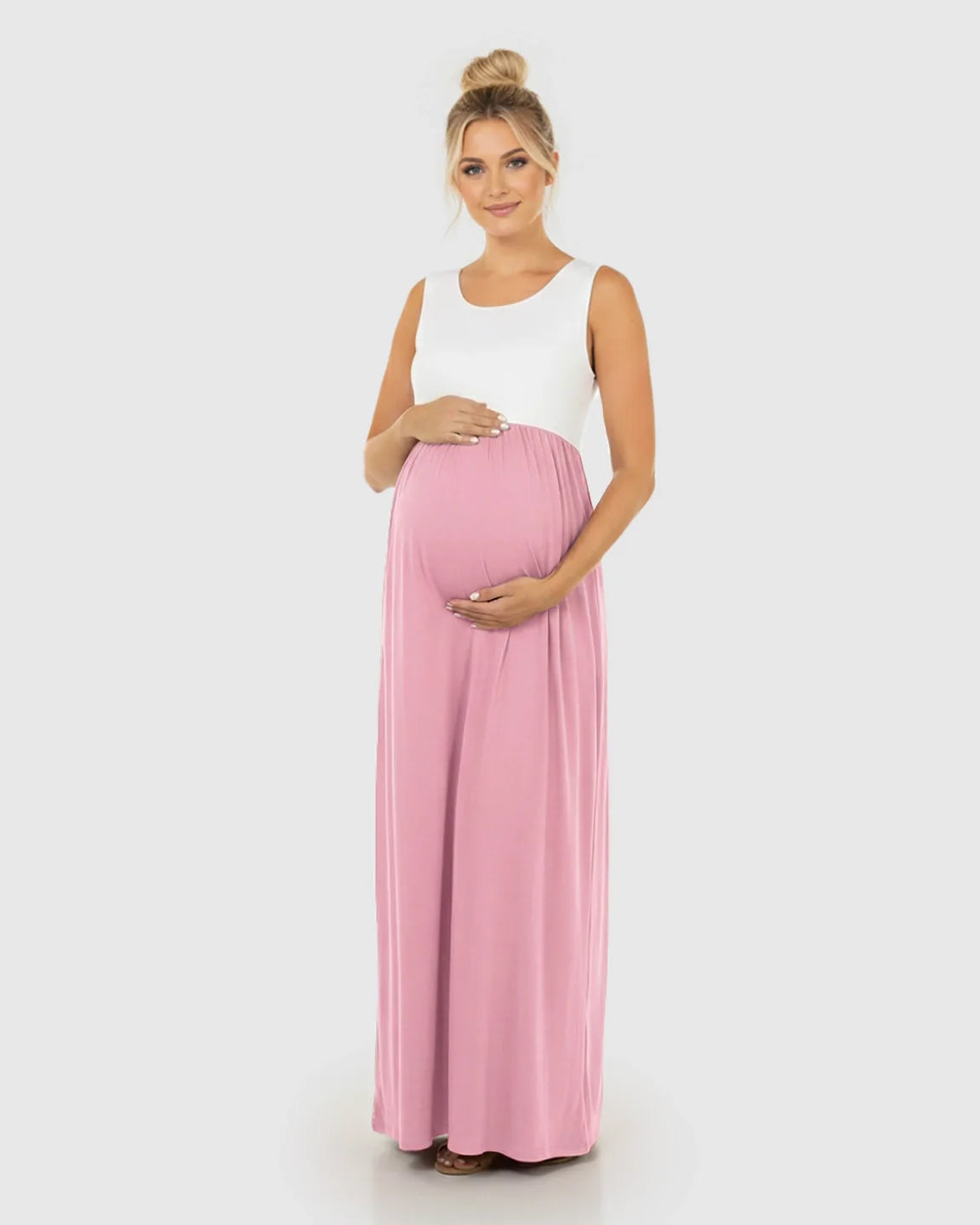 Maternity maxi dress: white sleeveless top, pink high-waisted flowing skirt. Pregnant woman cradles her bump.
