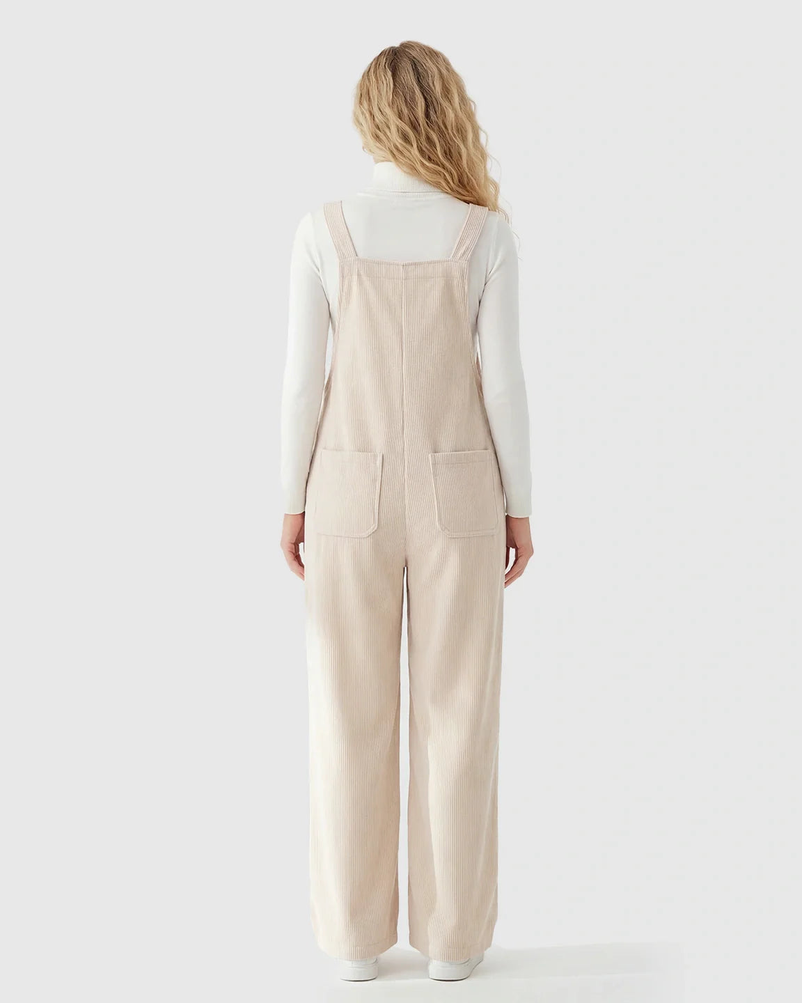 Blonde woman from behind in beige corduroy maternity overalls with adjustable straps and wide legs over a white turtleneck.