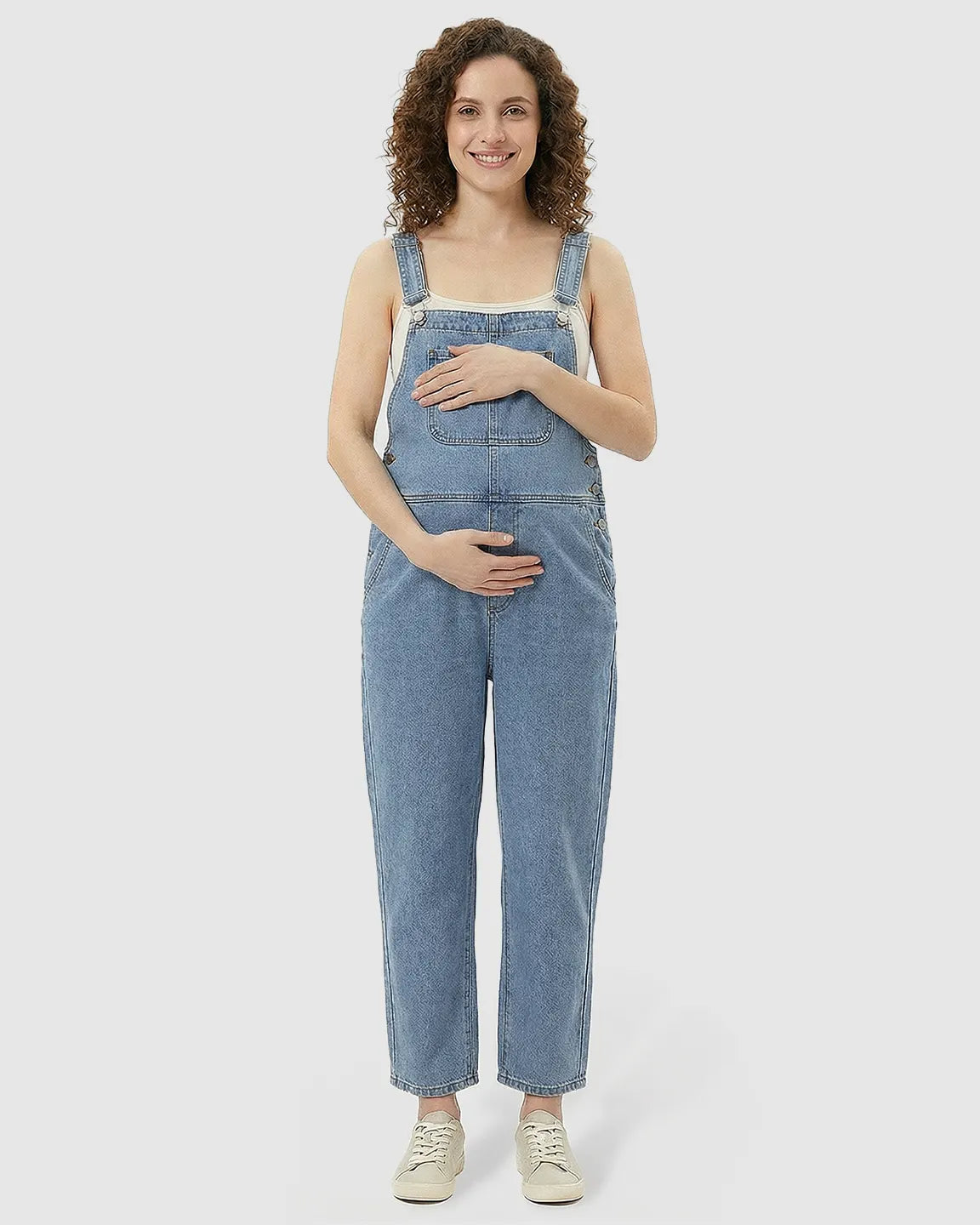 Smiling pregnant woman in a light wash loose denim maternity jumpsuit with adjustable straps.